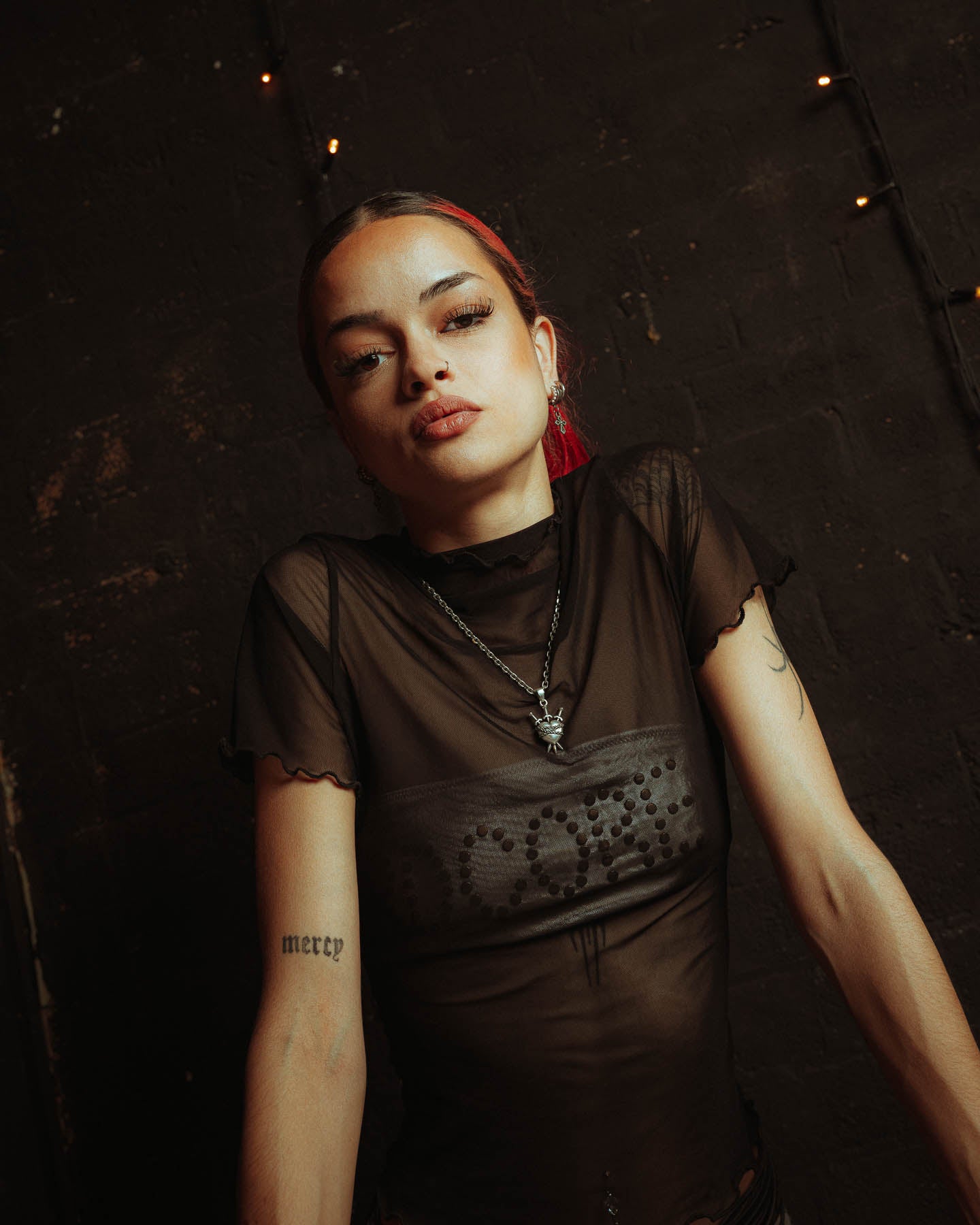 A person with red hair and facial piercings poses against a dark brick wall with string lights, wearing a sheer black top, a visible "mercy" tattoo on their left arm, and the 925 Sterling Silver "3 of Swords" Pendant.