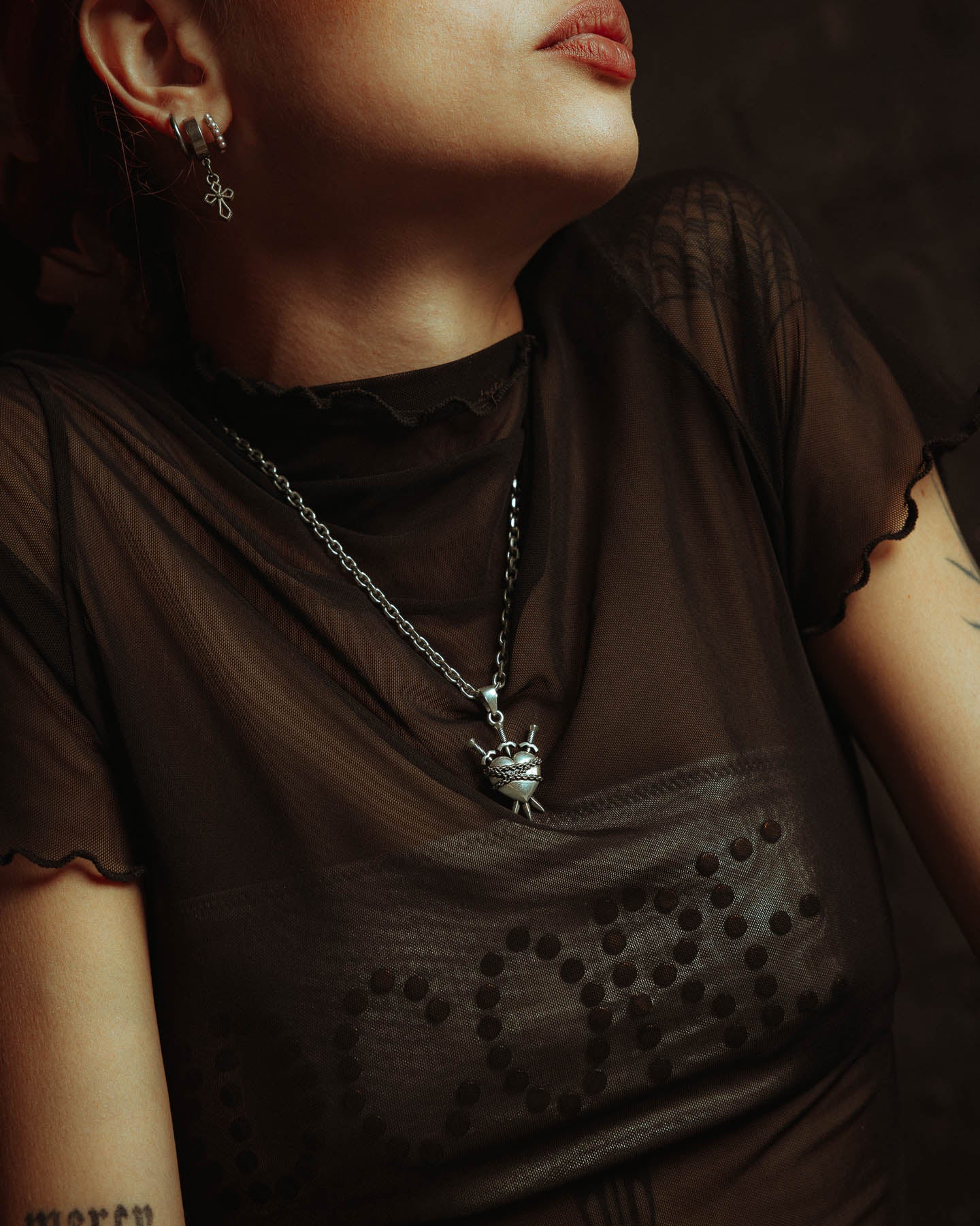 A person wearing a sheer black top with dotted lettering, a distinctive earring, tattoos on their partially turned arm, and the 925 Sterling Silver "3 of Swords" Pendant—a bold piece of tarot jewelry.