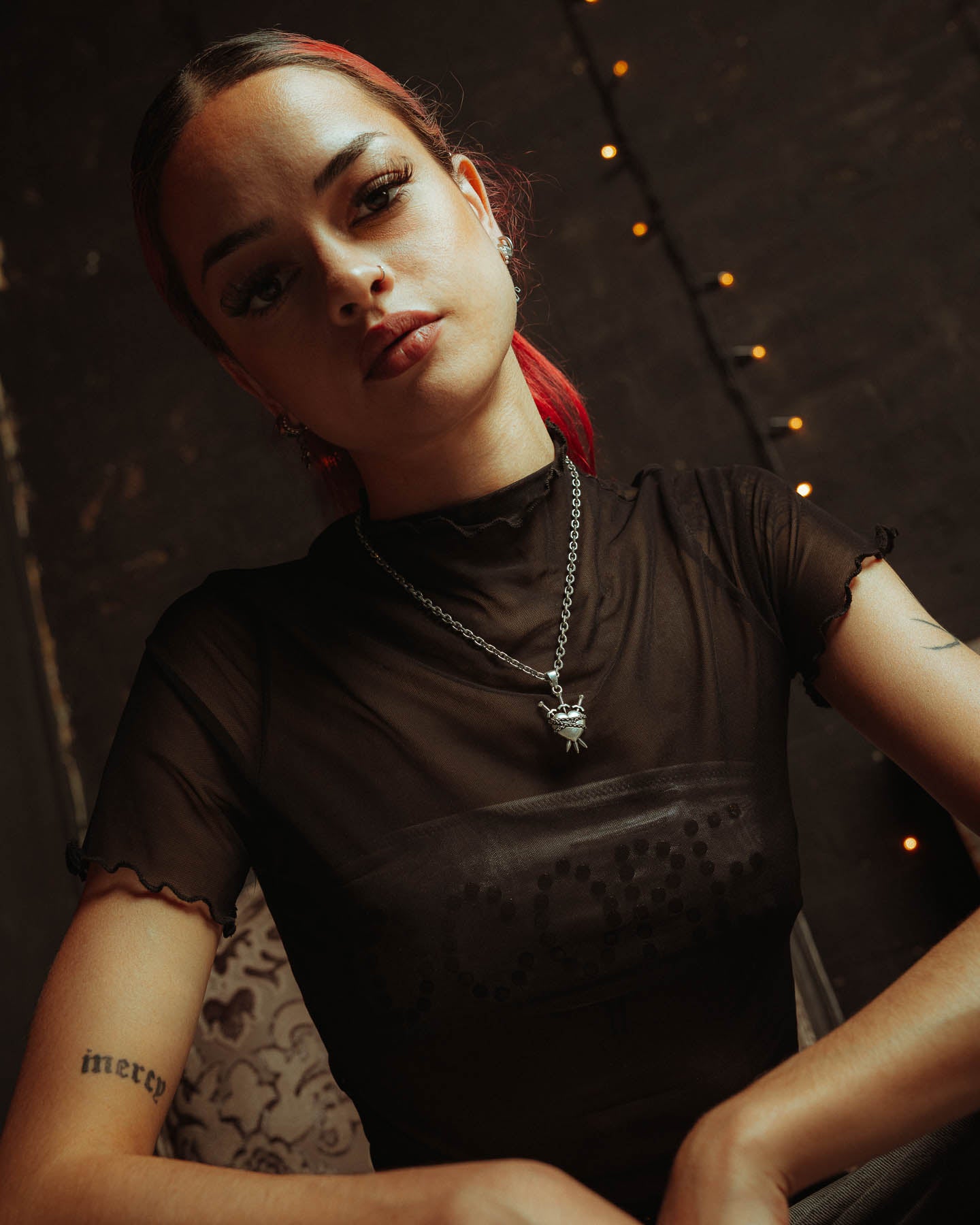 A person with red hair tied back wears a black sheer top, the 925 Sterling Silver "3 of Swords" Pendant necklace, and stud earrings. Tattoos cover their arms as they sit before a dim, string-lit backdrop, gazing confidently at the camera.