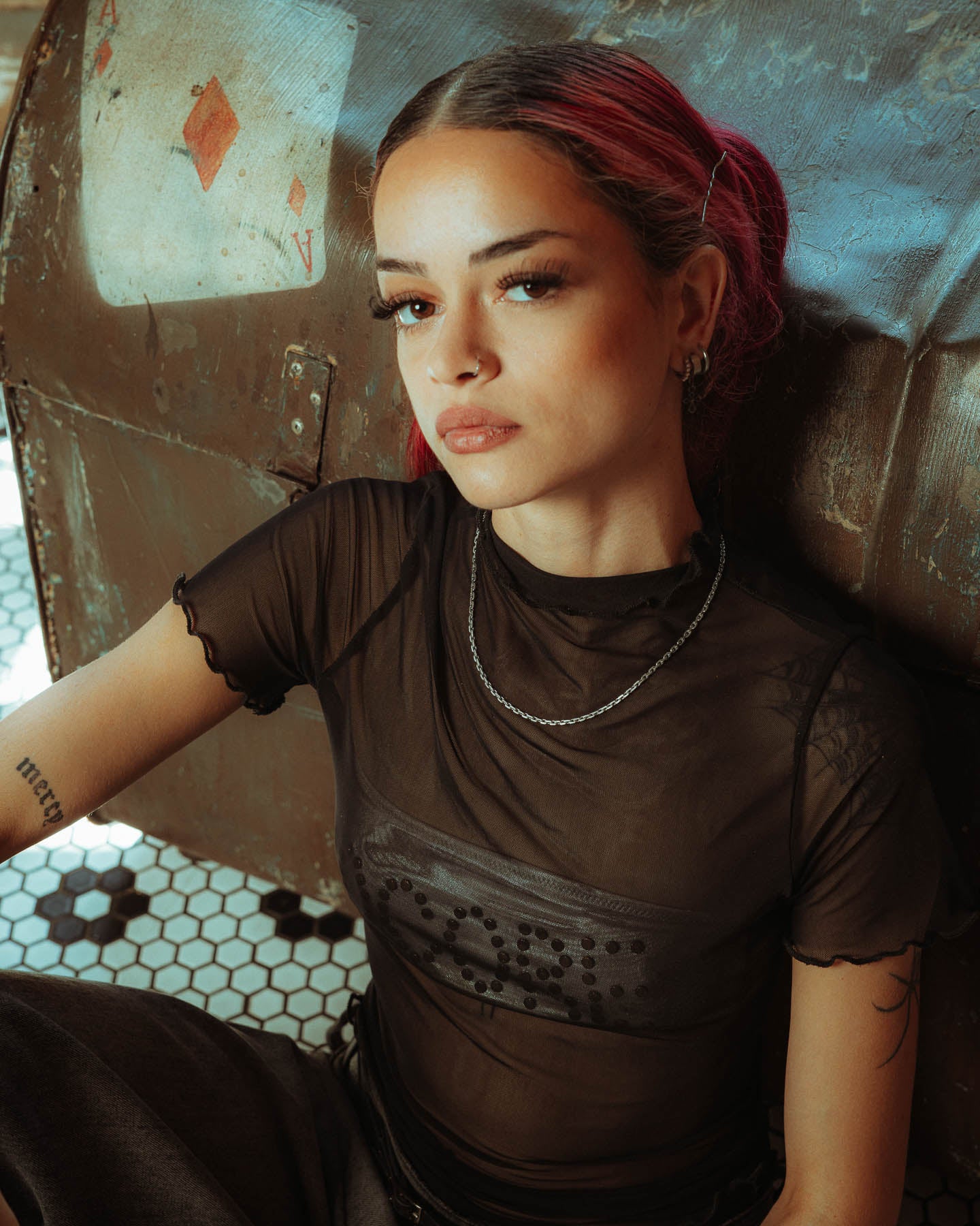 A young woman with pink hair and tattoos sits confidently against a metallic backdrop, wearing a sheer black top and a 925 Sterling Silver 3mm Cable Chain necklace.