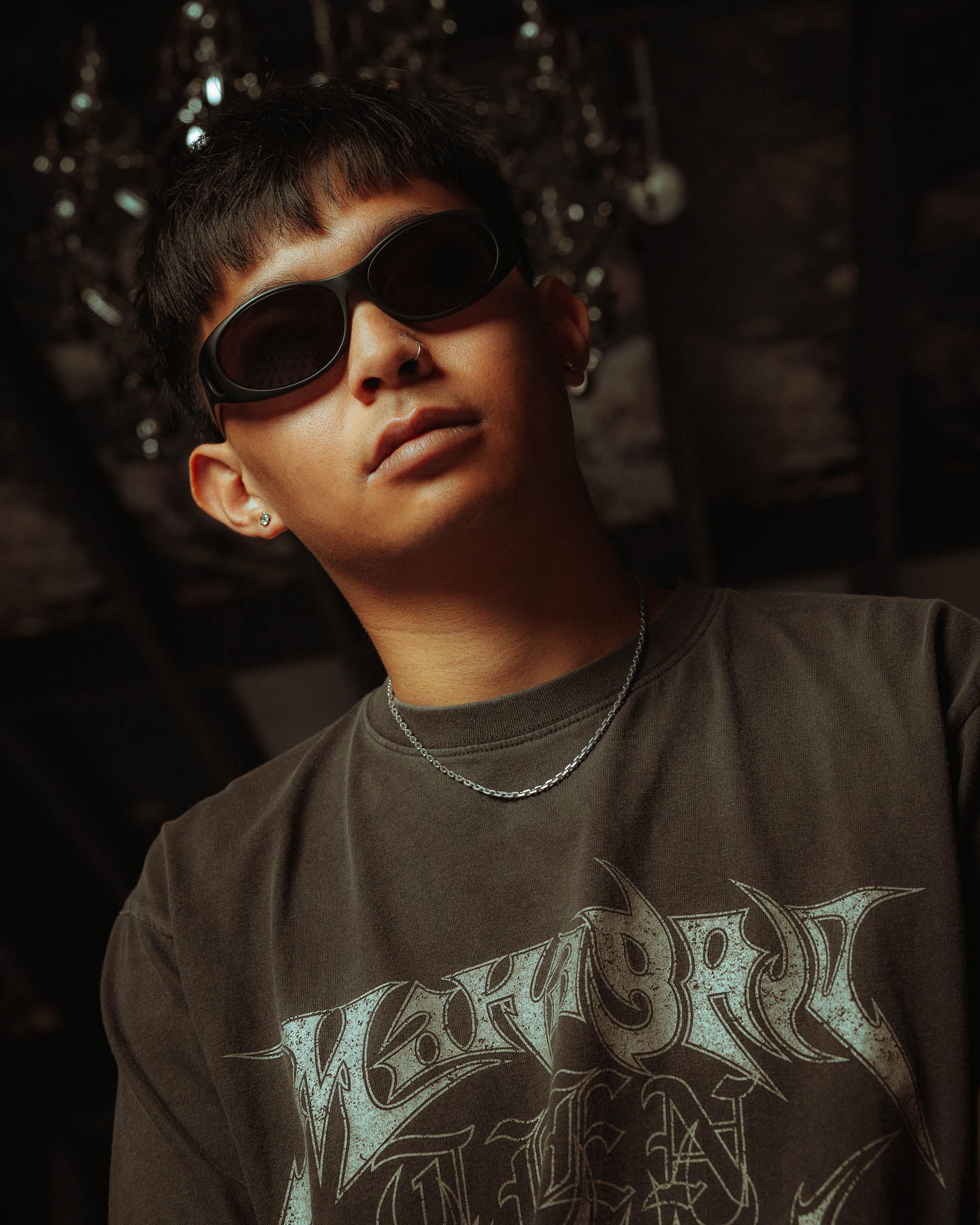 A young person in dark sunglasses and a graphic tee stands indoors with a serious expression, wearing the 925 Sterling Silver 3mm Cable Chain. The dim background features a chandelier and wooden beams above.