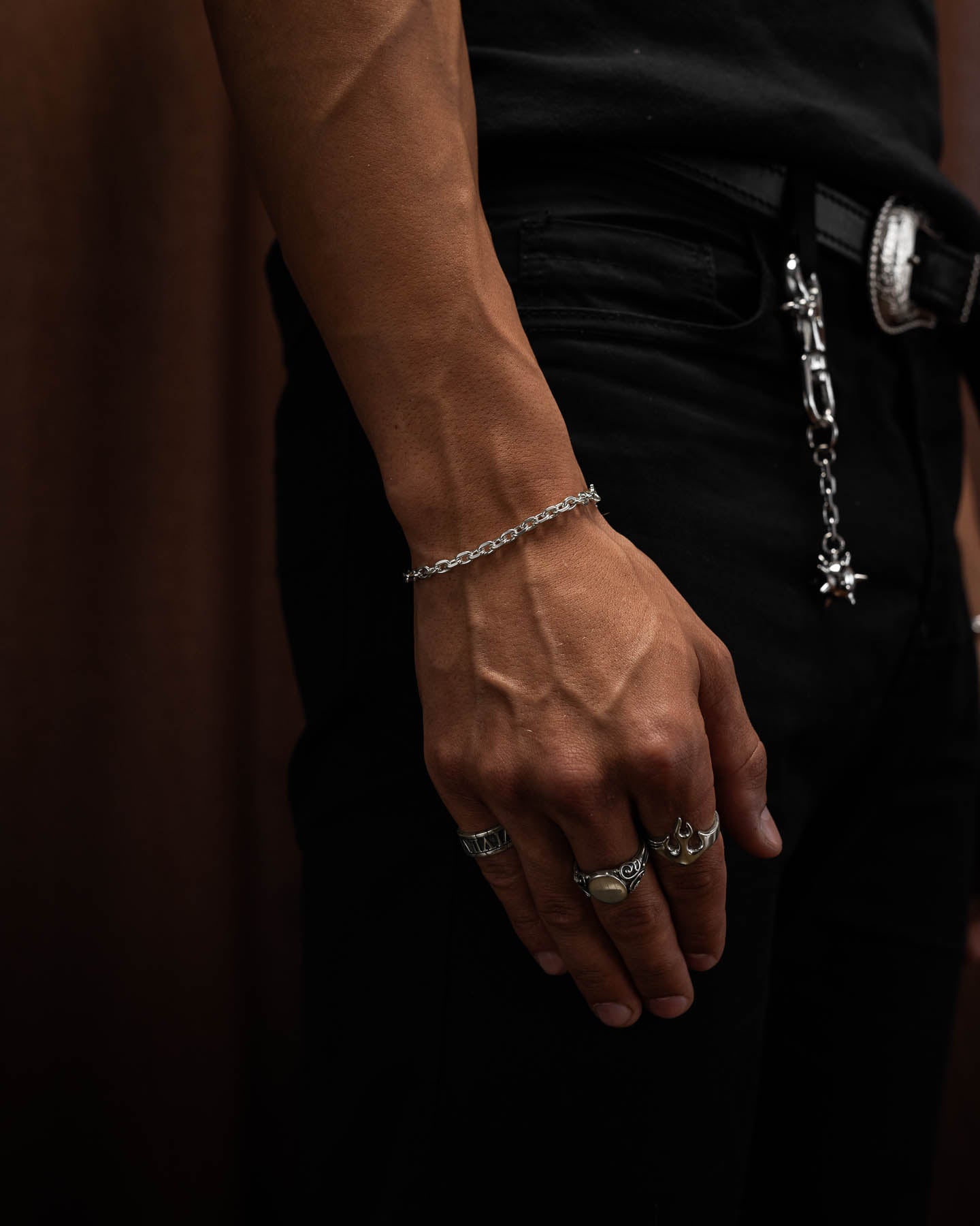 male model in tight black compression top holding first up to show the 4.5mm cable bracelet in sterling silver around his wrist with hand flat against leg to show off flame ring, oval signet ring and roman numerals ring in sterling silver.