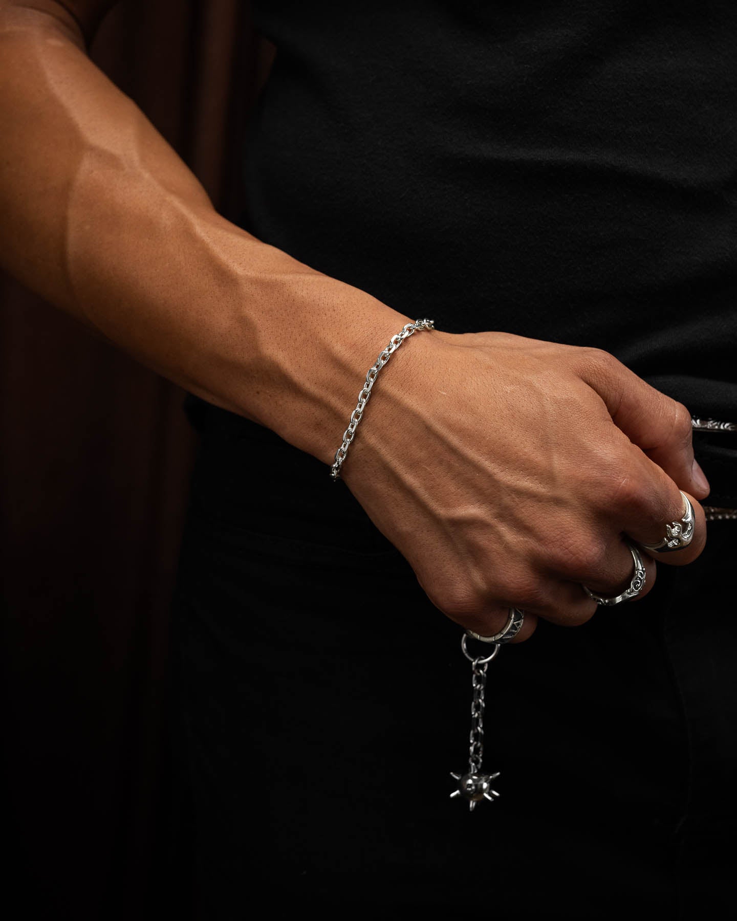 male model in tight black compression top holding first up to show the 4.5mm cable bracelet in sterling silver around his wrist. fist closed tensing with morning star spiked ball belt clip in the background