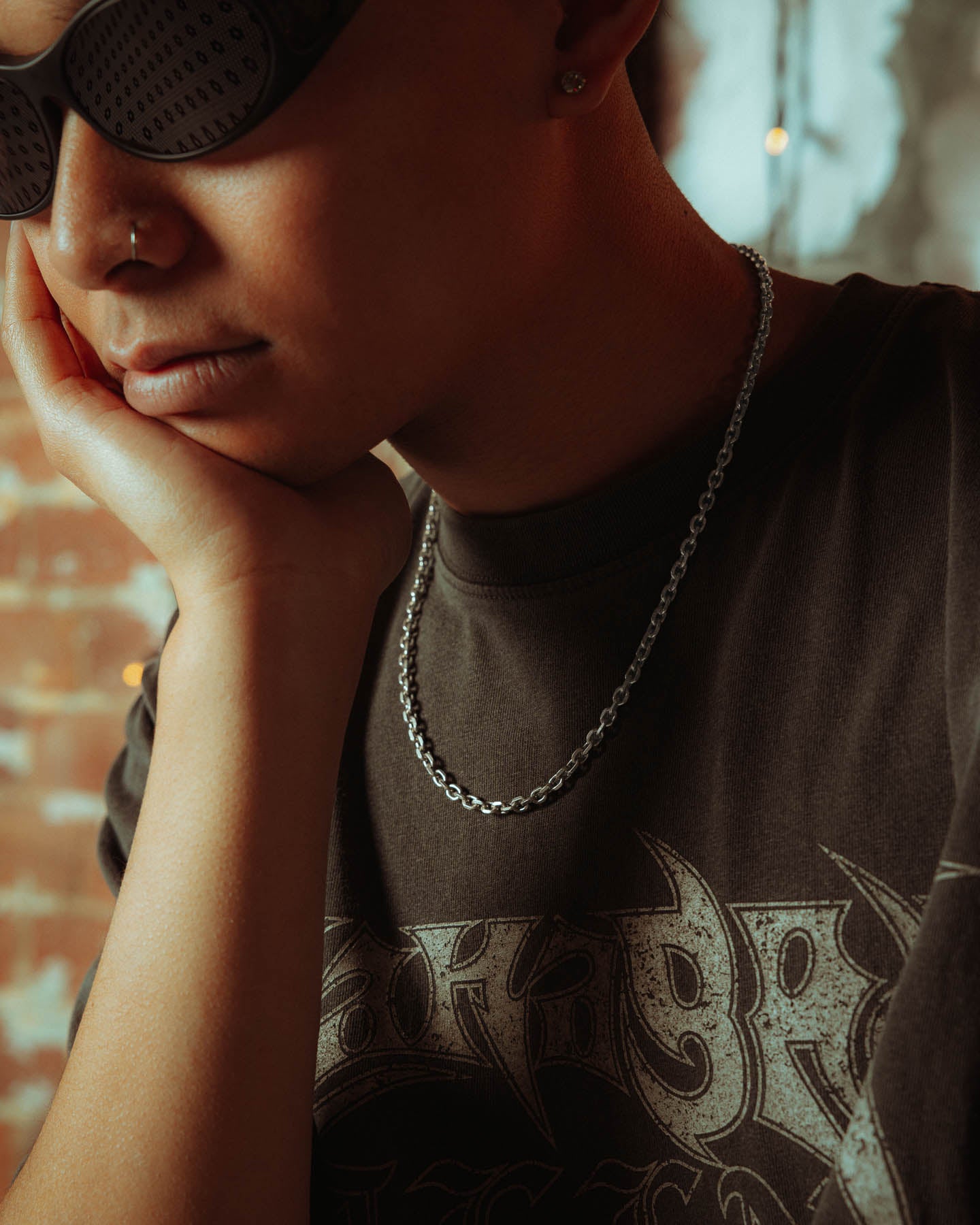 A person wearing dark sunglasses, a nose ring, and the 925 Sterling Silver 4.5mm Cable Chain rests their chin on their hand, sporting a black graphic tee with a brick wall and blurred hanging lights in the background.