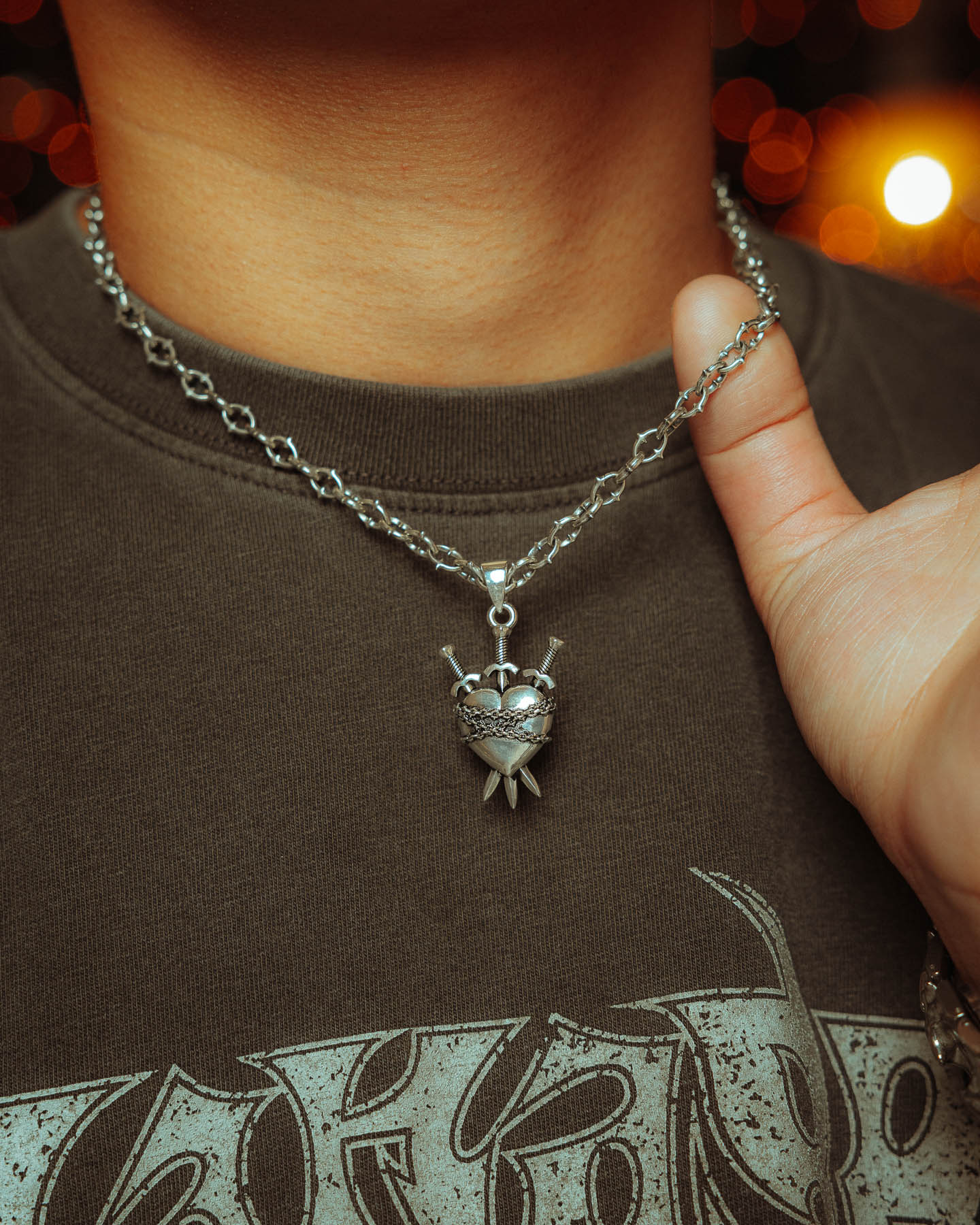 A person in a dark shirt holds "The Cathedral" 925 Sterling Silver 6mm Spiked Chain with a custom pendant, heart-shaped and pierced by three swords, against a backdrop of warm, glowing lights.
