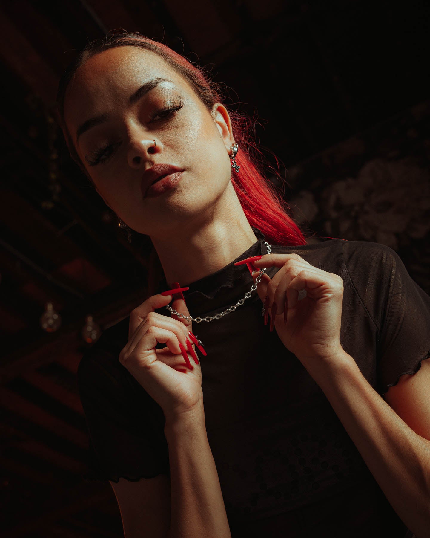 A woman with long red nails and tied-back red hair confidently poses in a sheer black top, holding the 925 Sterling Silver "The Cathedral" 6mm Spiked Chain against a dark, moody background.