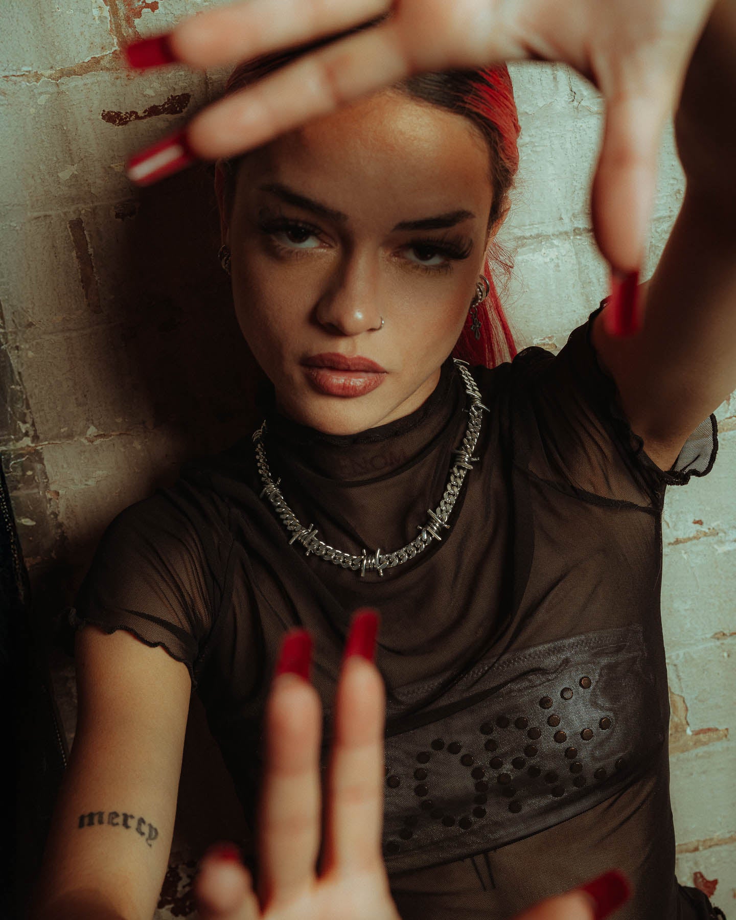 A person with red hair and long red nails poses near a brick wall, wearing a sheer black top, statement jewelry including a 925 Sterling Silver 9mm Barbed Wire Cuban Chain, a nose ring, and showing off their mercy tattoo.