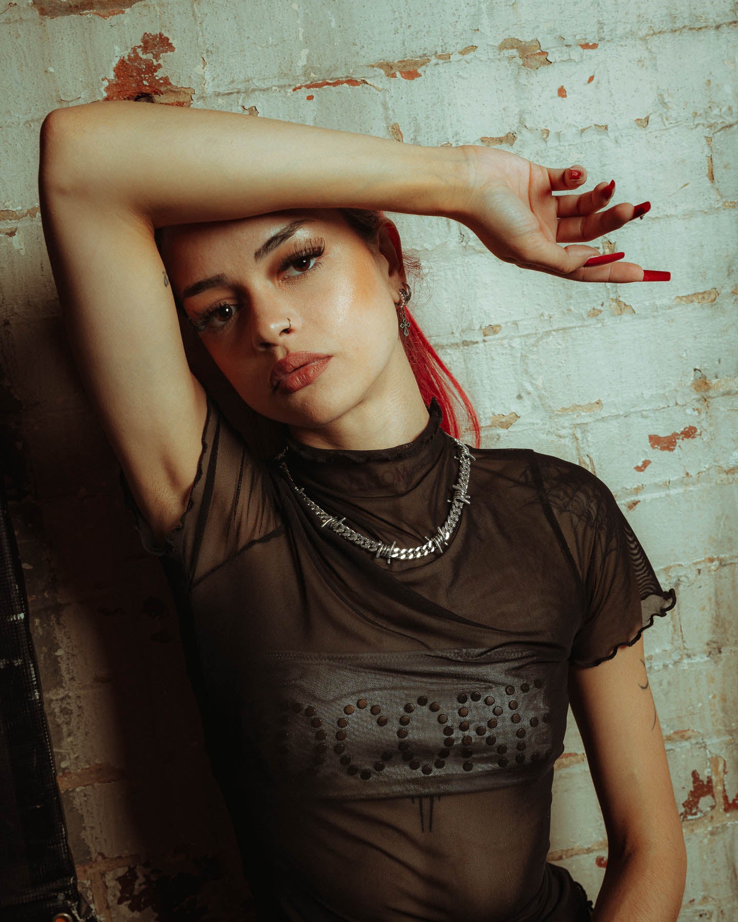 A woman with red hair and long red nails poses with one arm on her forehead, wearing a sheer black top and statement 925 Sterling Silver 9mm Barbed Wire Cuban Chain, standing against a textured white brick wall.