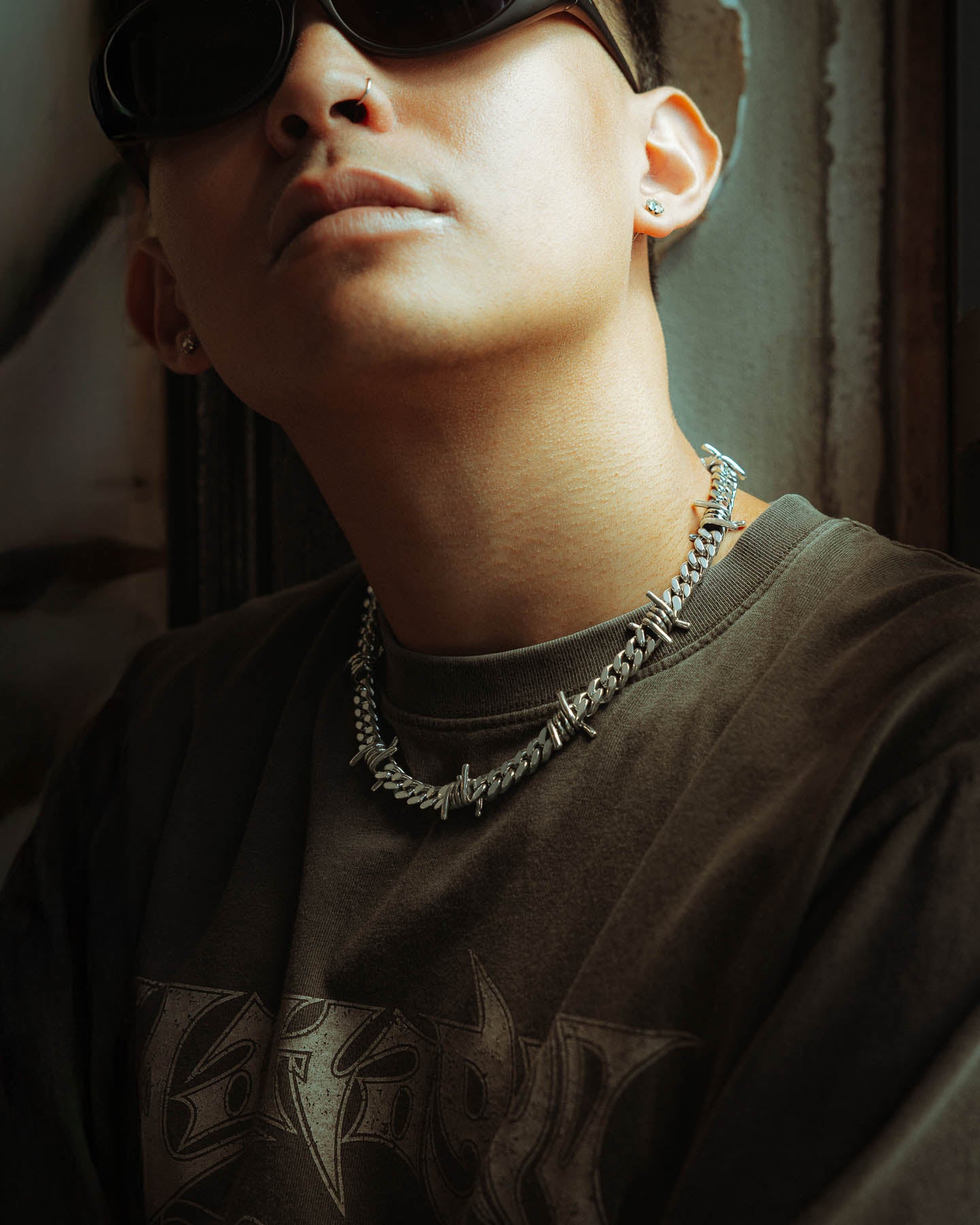 A person poses in moody lighting from a low angle, wearing dark sunglasses, a graphic t-shirt, and two bold jewelry pieces including the 925 Sterling Silver 9mm Barbed Wire Cuban Chain, with their partial face highlighted.
