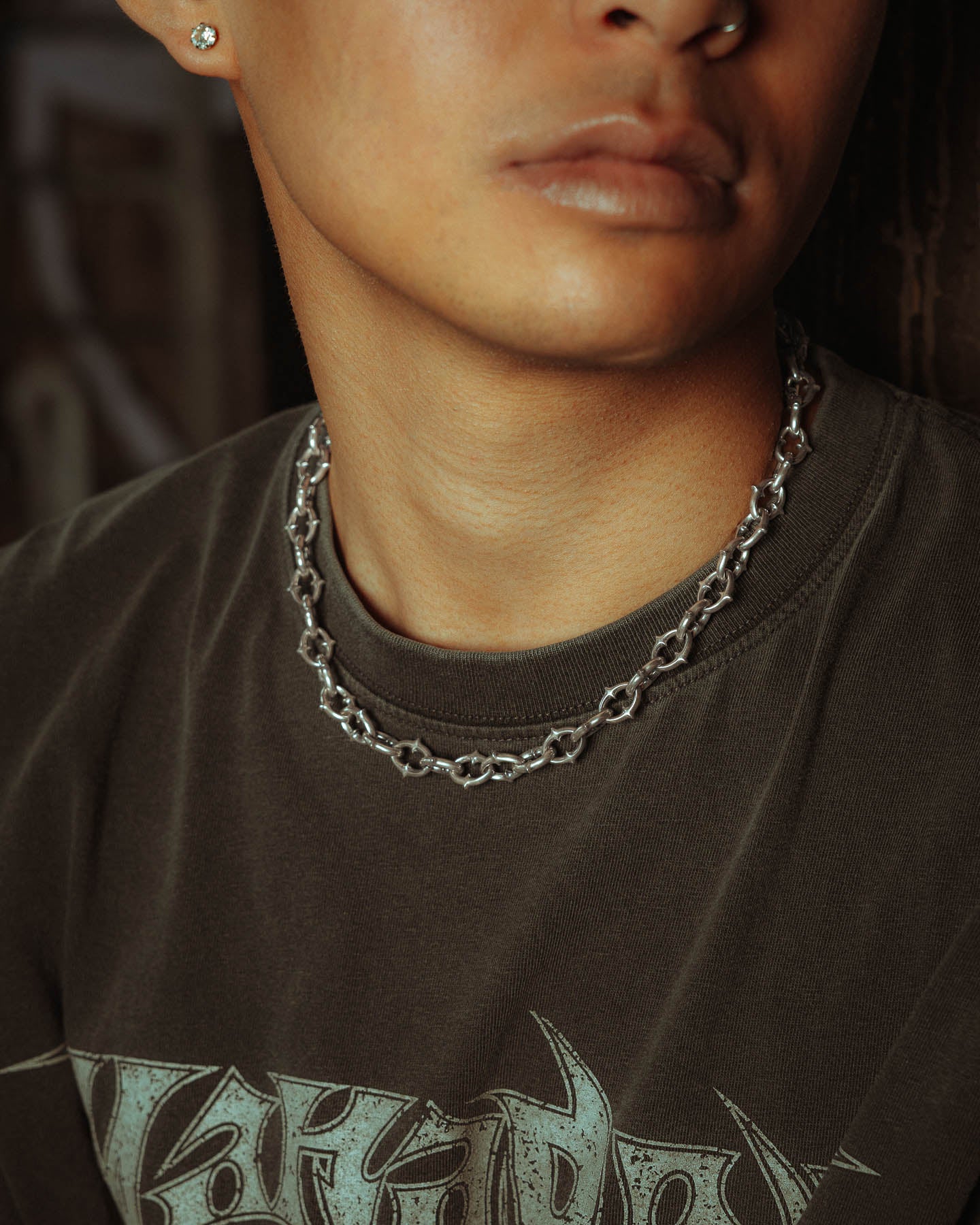 Close-up of a person wearing a dark graphic tee, a nose ring, stud earring, and "The Cathedral" 9mm Spiked Chain in 925 Sterling Silver. Only their lips and lower face are visible; eyes and forehead are out of frame.