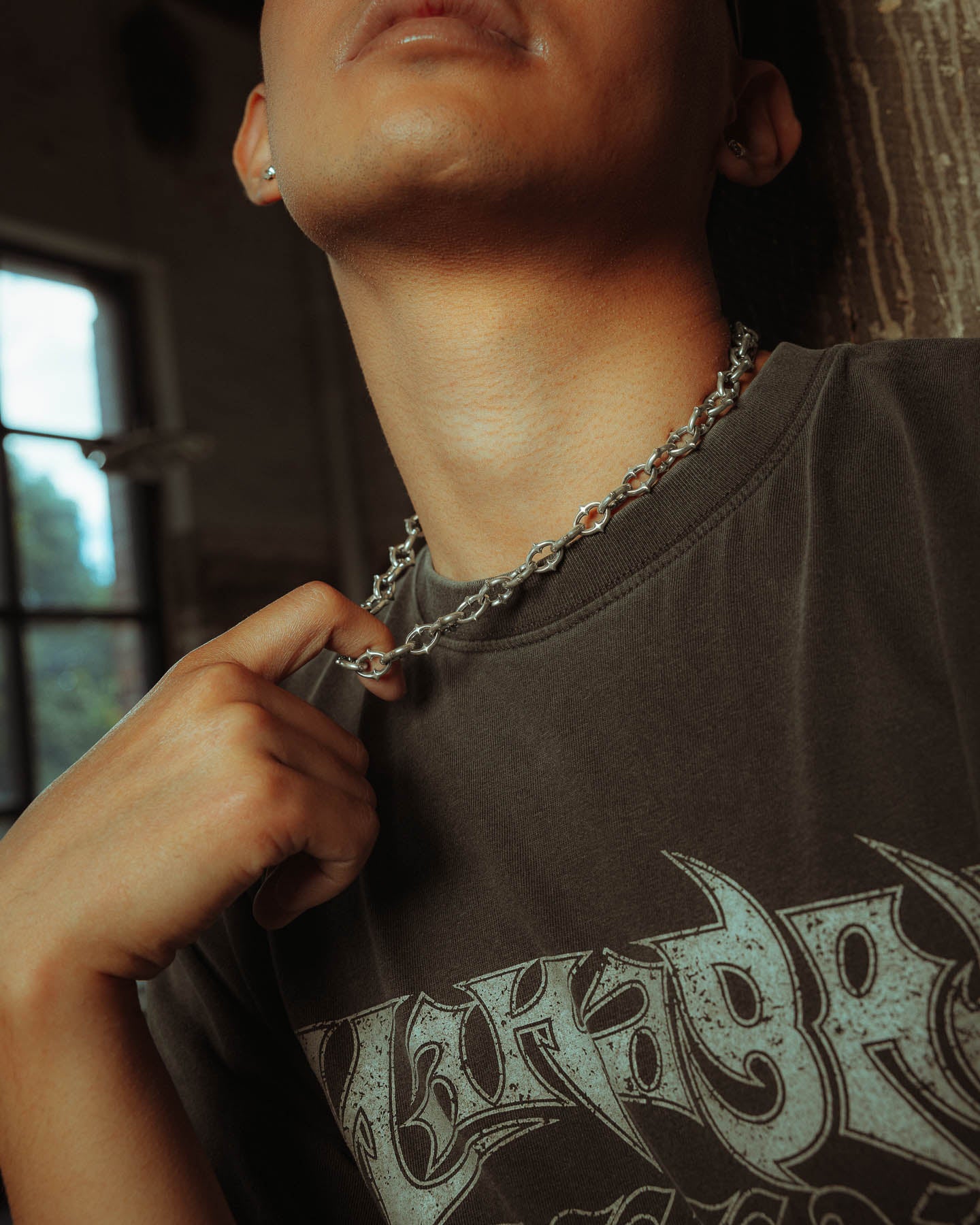 A person wearing a dark graphic t-shirt holds the 925 Sterling Silver "The Cathedral" 9mm Spiked Chain necklace against a backdrop with a window and wooden wall, the image cropped above their nose.