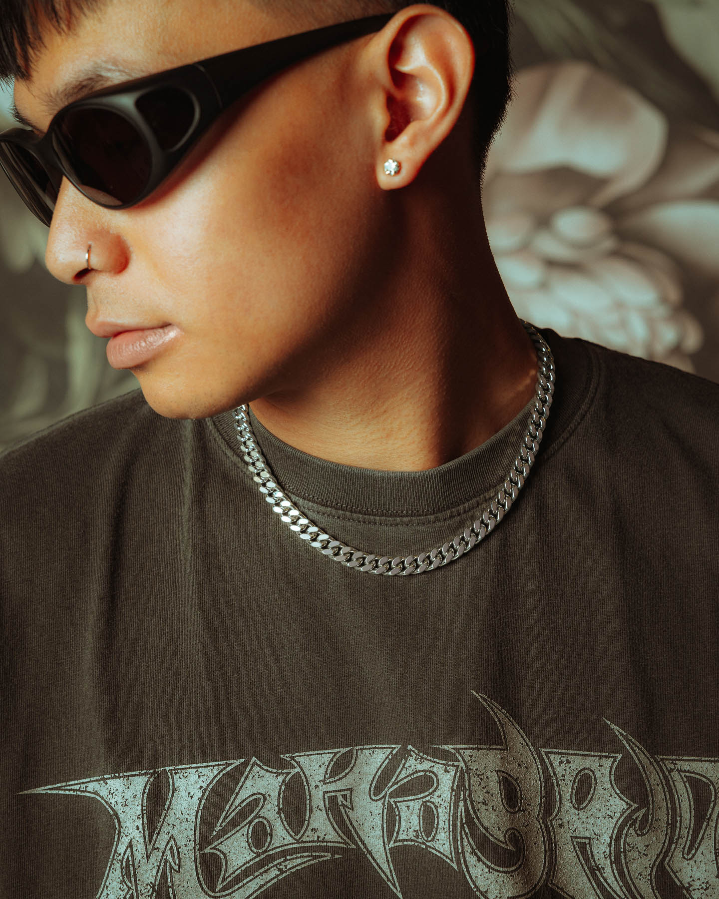 A person wearing dark sunglasses, a 925 Sterling Silver 9mm Cuban Chain, a nose ring, an earring, and a dark graphic t-shirt looks to the side against a floral-patterned background.