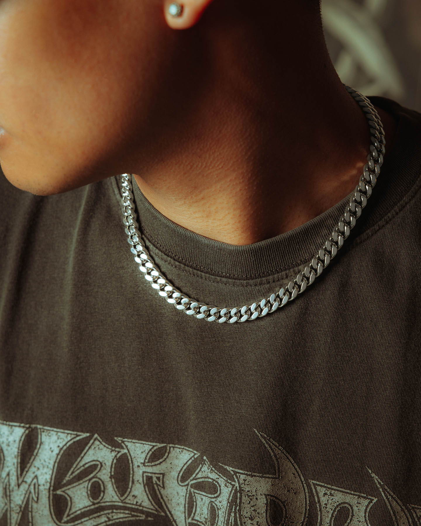 A person wears a 925 Sterling Silver 9mm Cuban Chain with a dark graphic t-shirt featuring metallic lettering. The bold silver chain stands out, paired with a small stud earring in one ear.