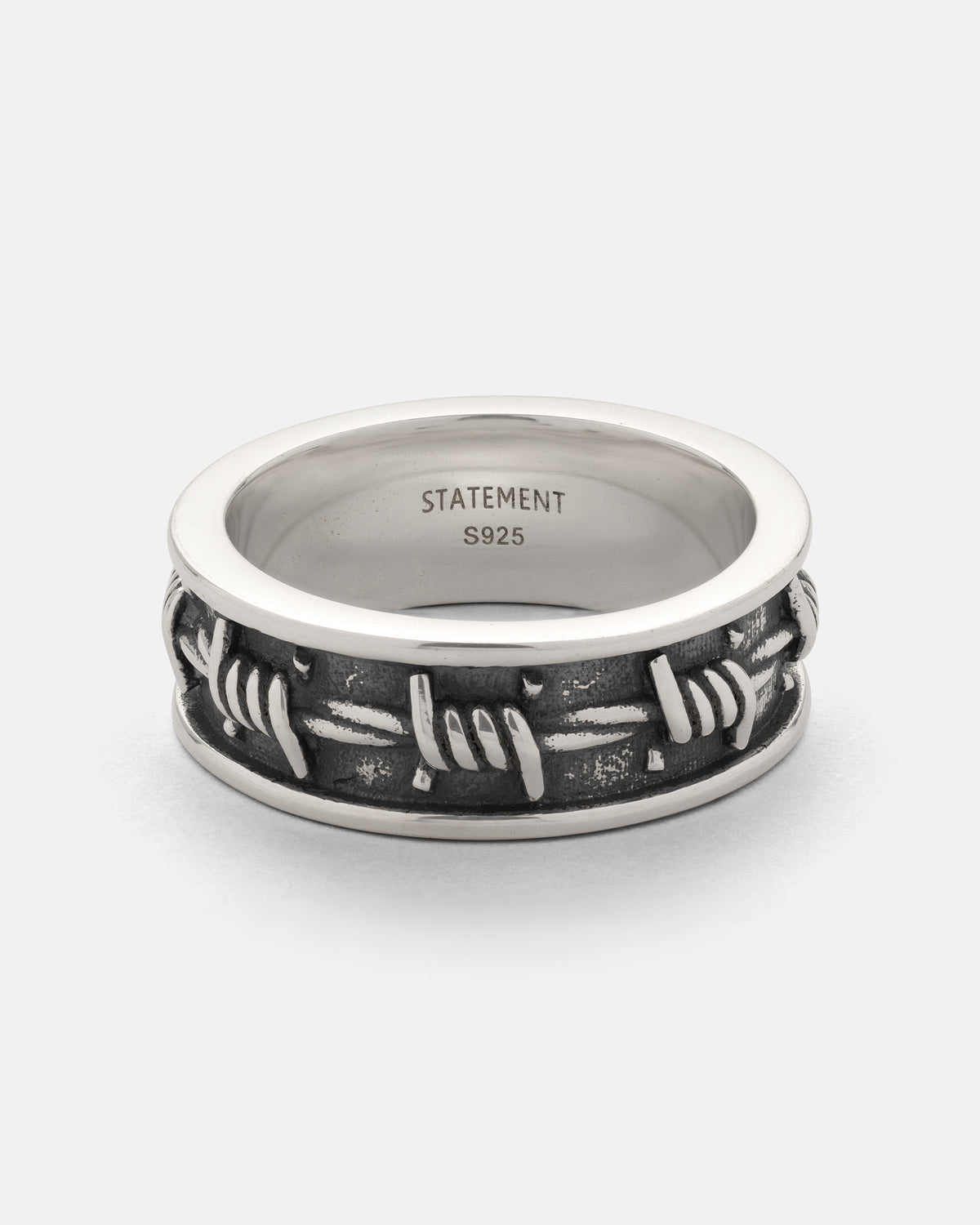 Introducing the 925 Sterling Silver Barbed Wire Ring by Statement Collective. This striking stackable ring features a raised, 3D barbed wire band that wraps around a polished sterling silver edge, set against a dark, textured background for eye-catching contrast. Inside the band, “STATEMENT S925” is finely engraved—a bold accessory with modern style and meticulous detail.