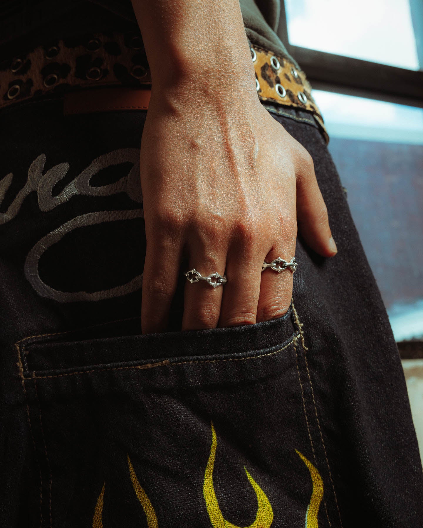 A hand wearing the 925 Sterling Silver "The Cathedral" Spiked Ring rests on the back pocket of black pants with yellow flame designs, visible stitching, and a metal eyelet belt.