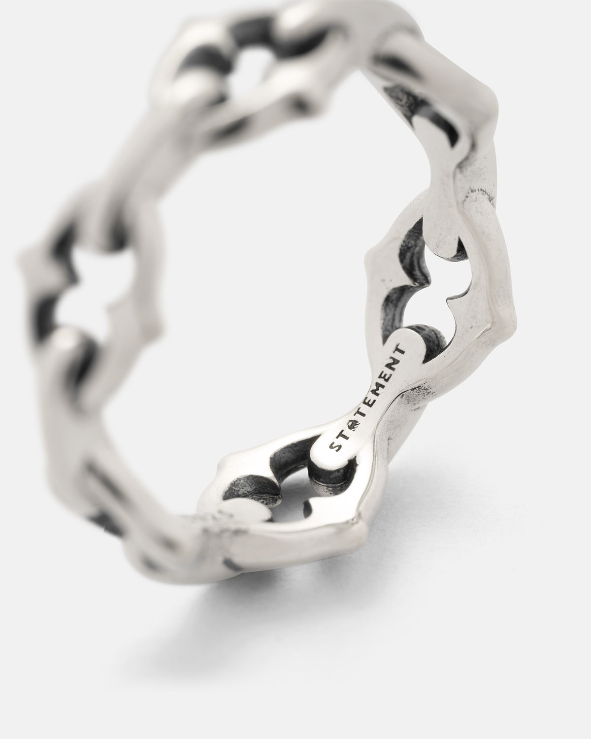 Introducing the 925 Sterling Silver “The Cathedral” Spiked Ring by Statement Collective. This striking ring features a unique interconnected design inspired by chain links, accented with subtle dark details. The inner band is engraved with the word “STATEMENT.” Presented against a plain white background.