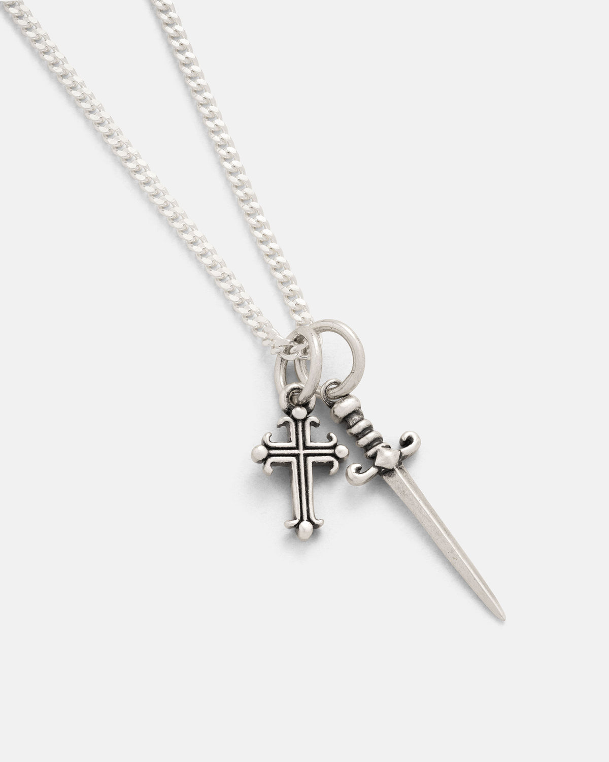 The Statement Collective 925 Sterling Silver Cross & Dagger Necklace showcases a sleek silver chain adorned with two pendants—a small, rounded-tip cross and a finely detailed dagger—suspended side by side against a plain backdrop. This bold piece of rock jewelry is highlighted by its intricate design elements, making it an eye-catching sterling silver pendant set ideal for any collection.