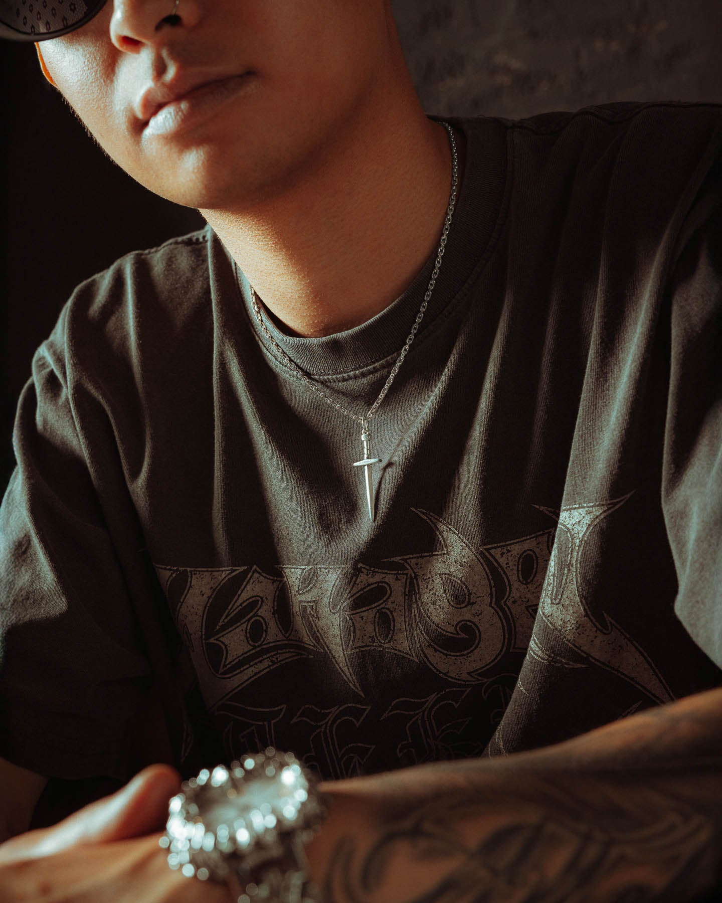A person wears a dark graphic t-shirt, a cross necklace, and a 925 Sterling Silver Dagger Pendant. Their tattooed arm is visible, and their face is partially out of frame and lit by soft light.
