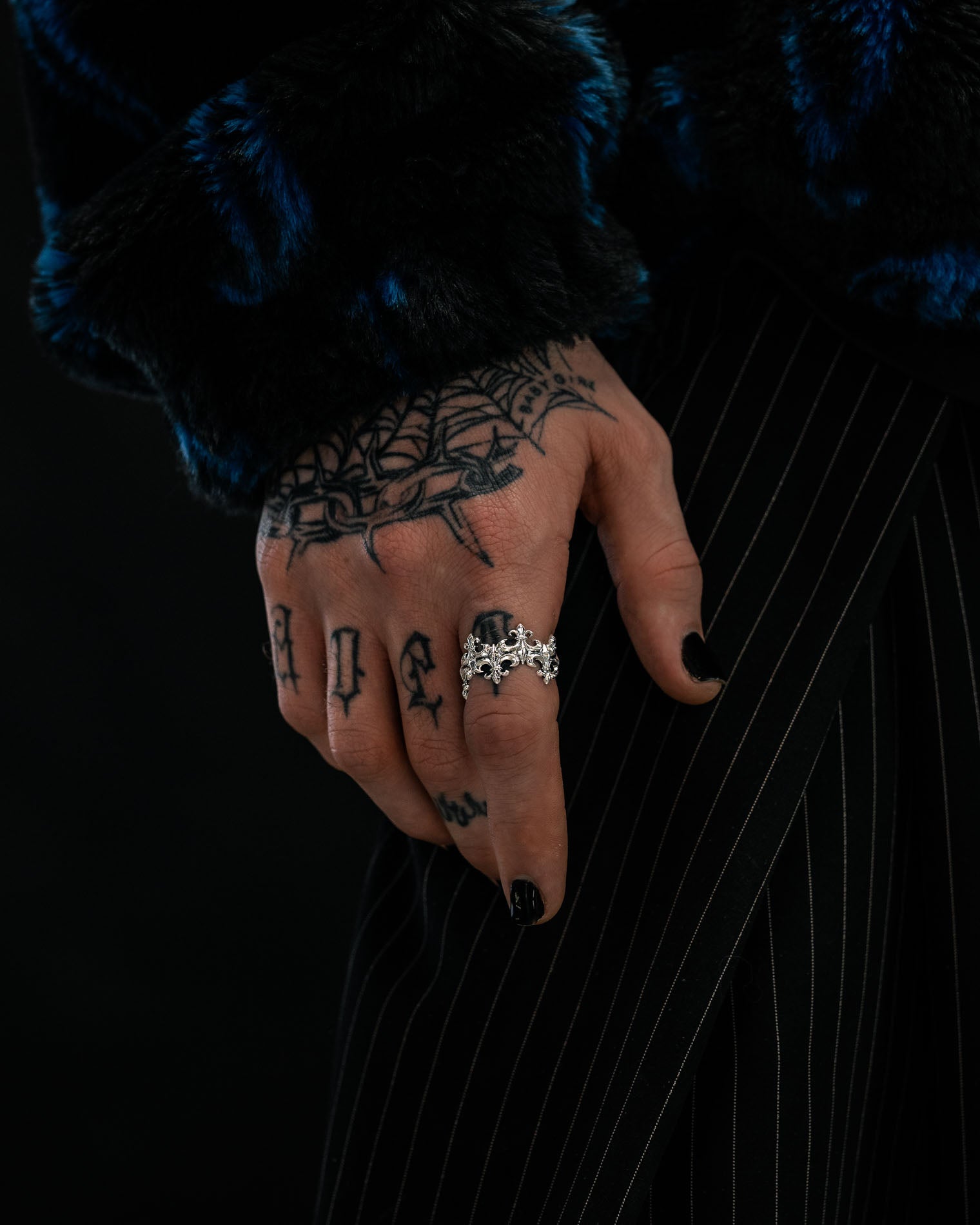 A tattooed hand with black nail polish wears the Statement Collective 925 Sterling Silver “Crested” Fleur De Lis Ring. The person is dressed in a blue and black fur coat and black pinstripe pants, showcasing their love for vintage silver jewelry.