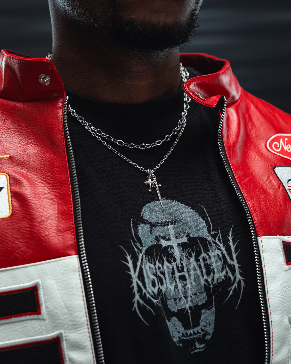 A person wearing a red and white jacket, a black graphic t-shirt with a screaming skull design, and two silver chain necklaces—including the Statement Collective 925 Sterling Silver Cross & Dagger Necklace—with bold rock-inspired jewelry. The face is partially out of frame.