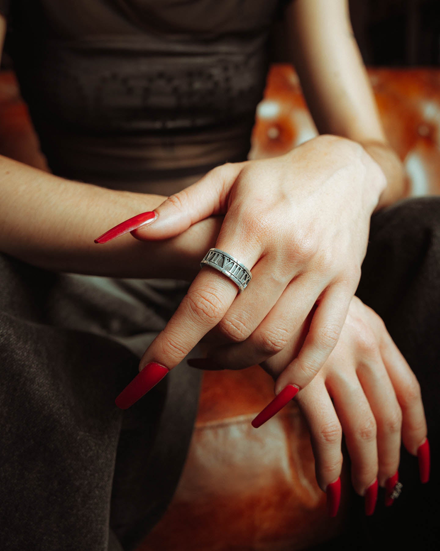 A person with long, red, pointed nails rests their hands together, showcasing the 925 Sterling Silver Numerals Ring. They are seated on a brown leather couch, dressed in dark pants and a sheer black top.