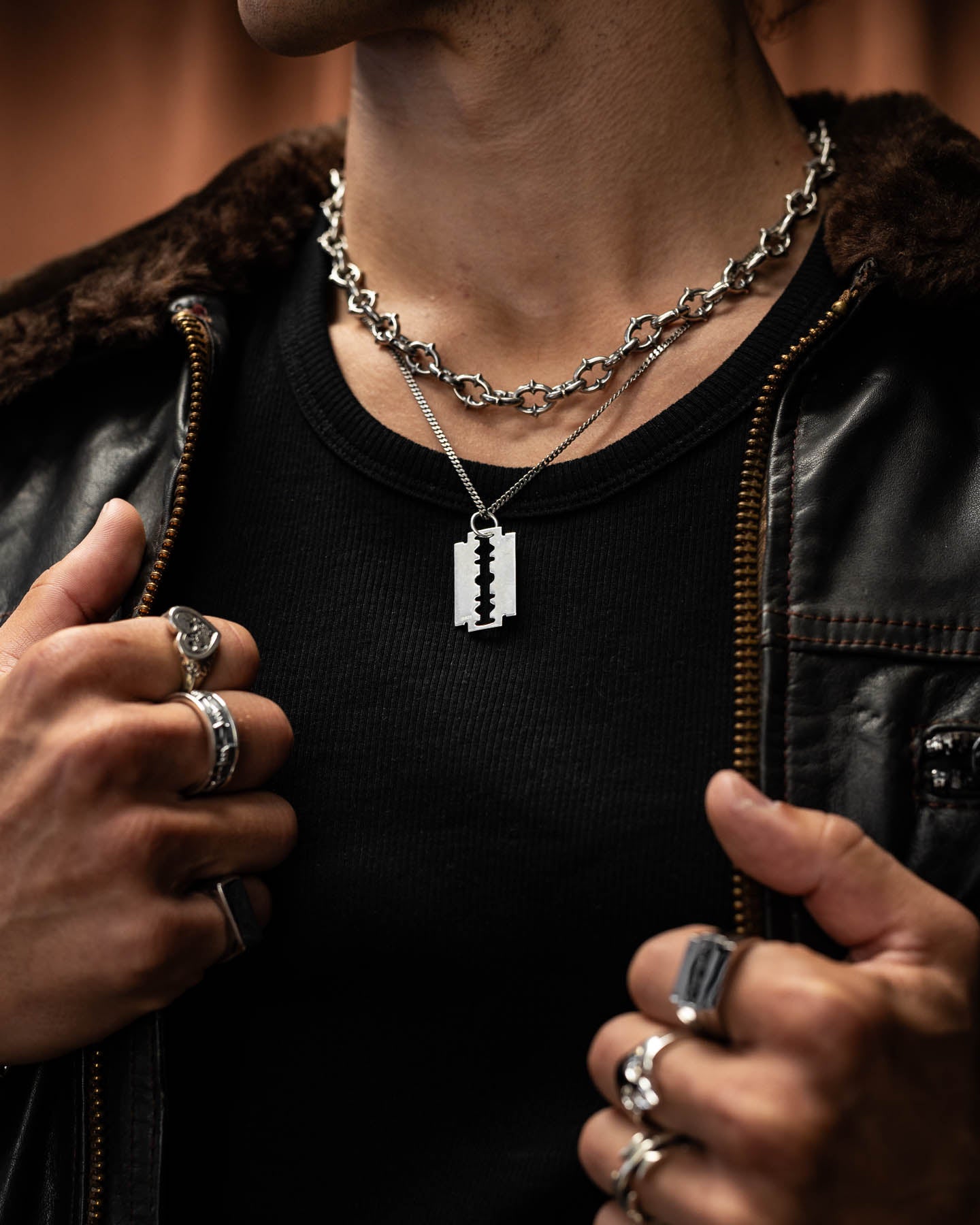A person in punk metal style—silver rings, black shirt, leather jacket—shows off two necklaces: a chunky chain and the 925 Sterling Silver Razor Blade Pendant, holding their jacket open.