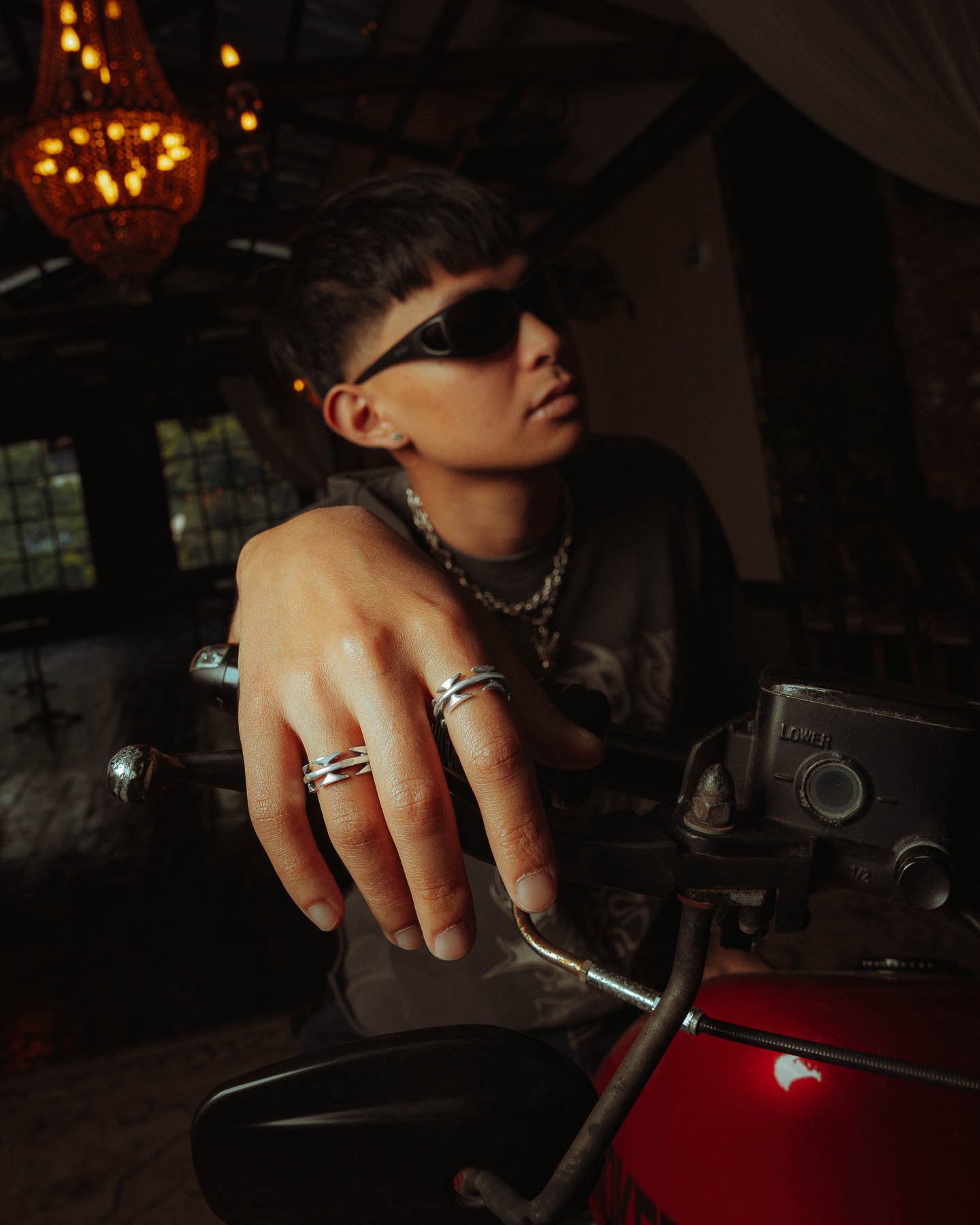 A person wearing sunglasses and a 925 Sterling Silver Razor Wire Ring sits on a red motorcycle indoors, gripping the handlebar, with a chandelier and large windows visible in the dimly lit background.