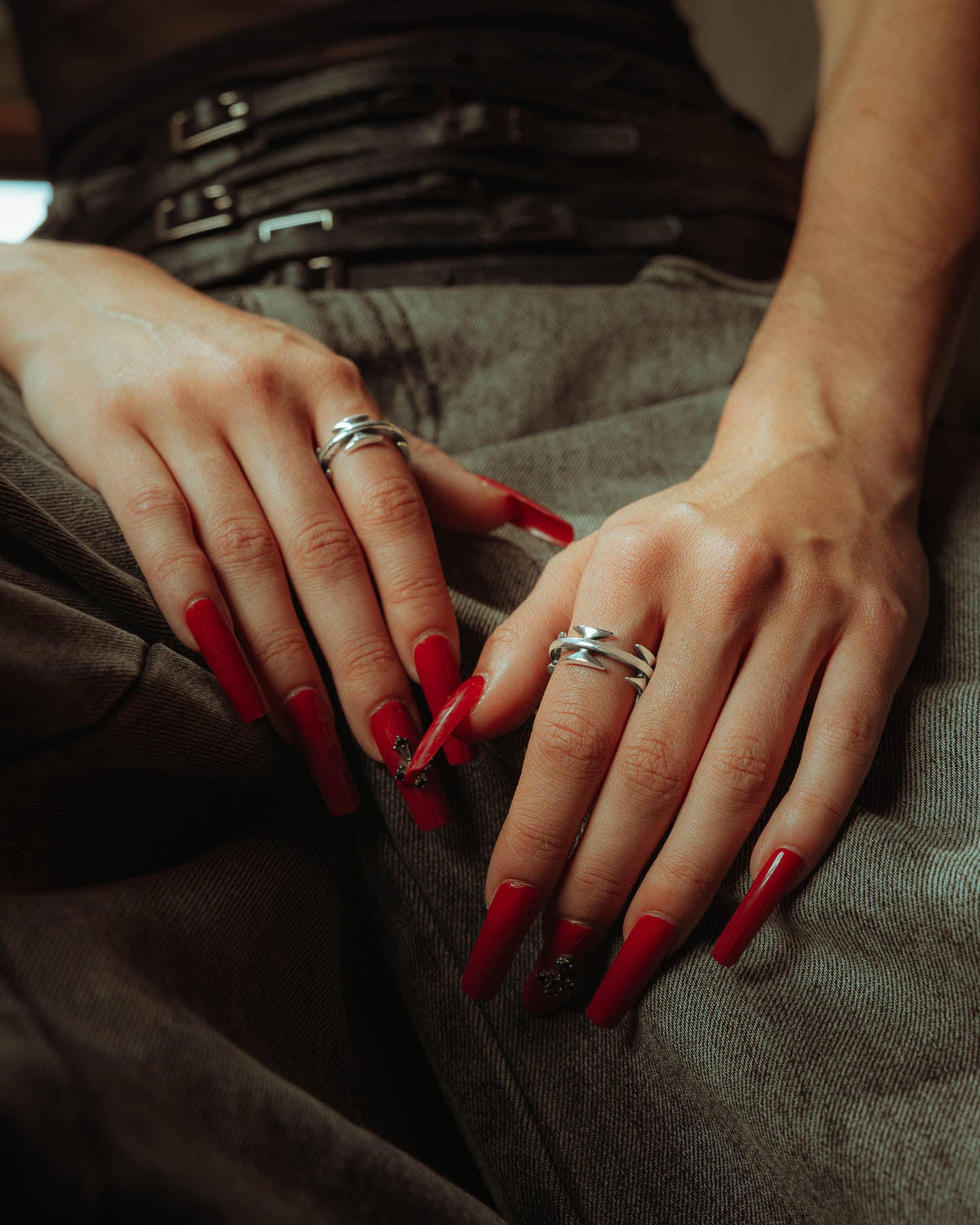 Close-up of two hands with long red nails resting on denim jeans, wearing multiple rings including the 925 Sterling Silver Razor Wire Ring. A black belt is visible at the waist, with soft lighting enhancing the tattoo-inspired jewelry.