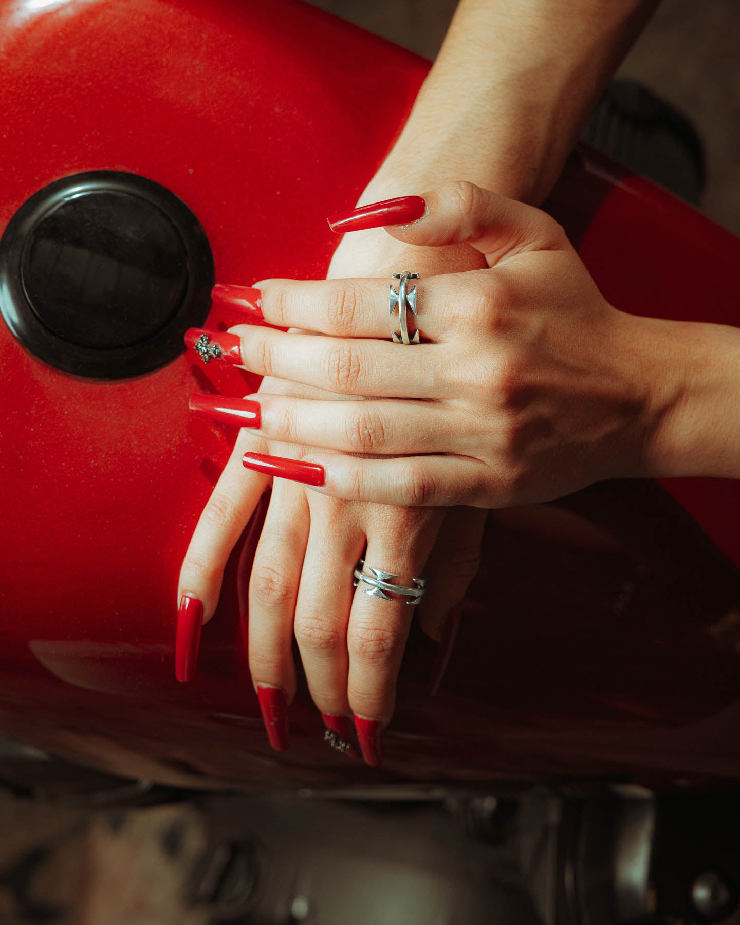 Two hands with long, red nails and several sterling silver rings—including the 925 Sterling Silver Razor Wire Ring—rest on a shiny red surface, possibly a motorcycle’s fuel tank. One hand gently holds the other.