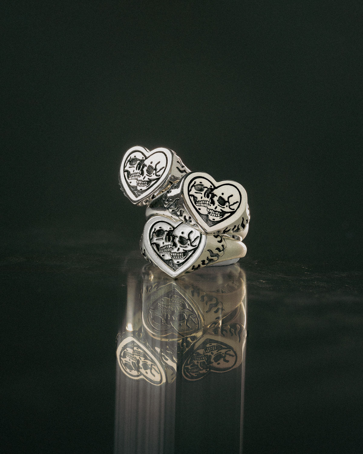 Three 925 Sterling Silver Skull Heart Rings are stacked on a reflective dark surface, each featuring an engraved skull and crossbones on a heart-shaped face, highlighting the bold style of handcrafted silver jewelry.