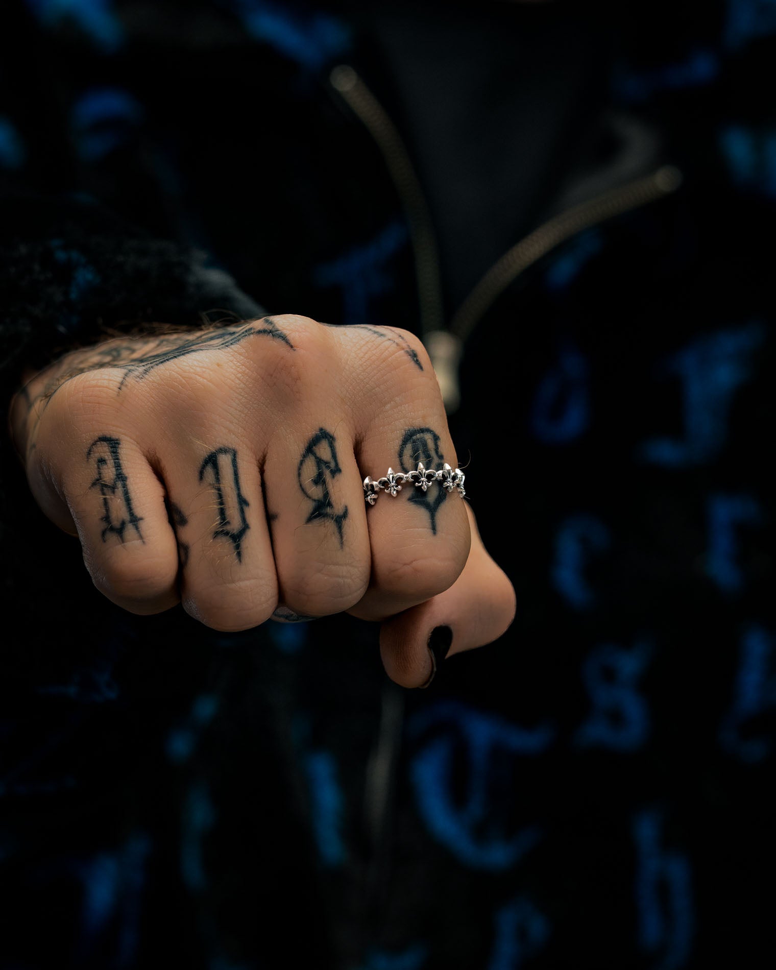 A close-up of a fist with black letter tattoos on the fingers, showcasing the Statement Collective 925 Sterling Silver “Fleur De Lis” Ring worn on the middle finger. The person wears a dark, patterned jacket adorned with blue designs.