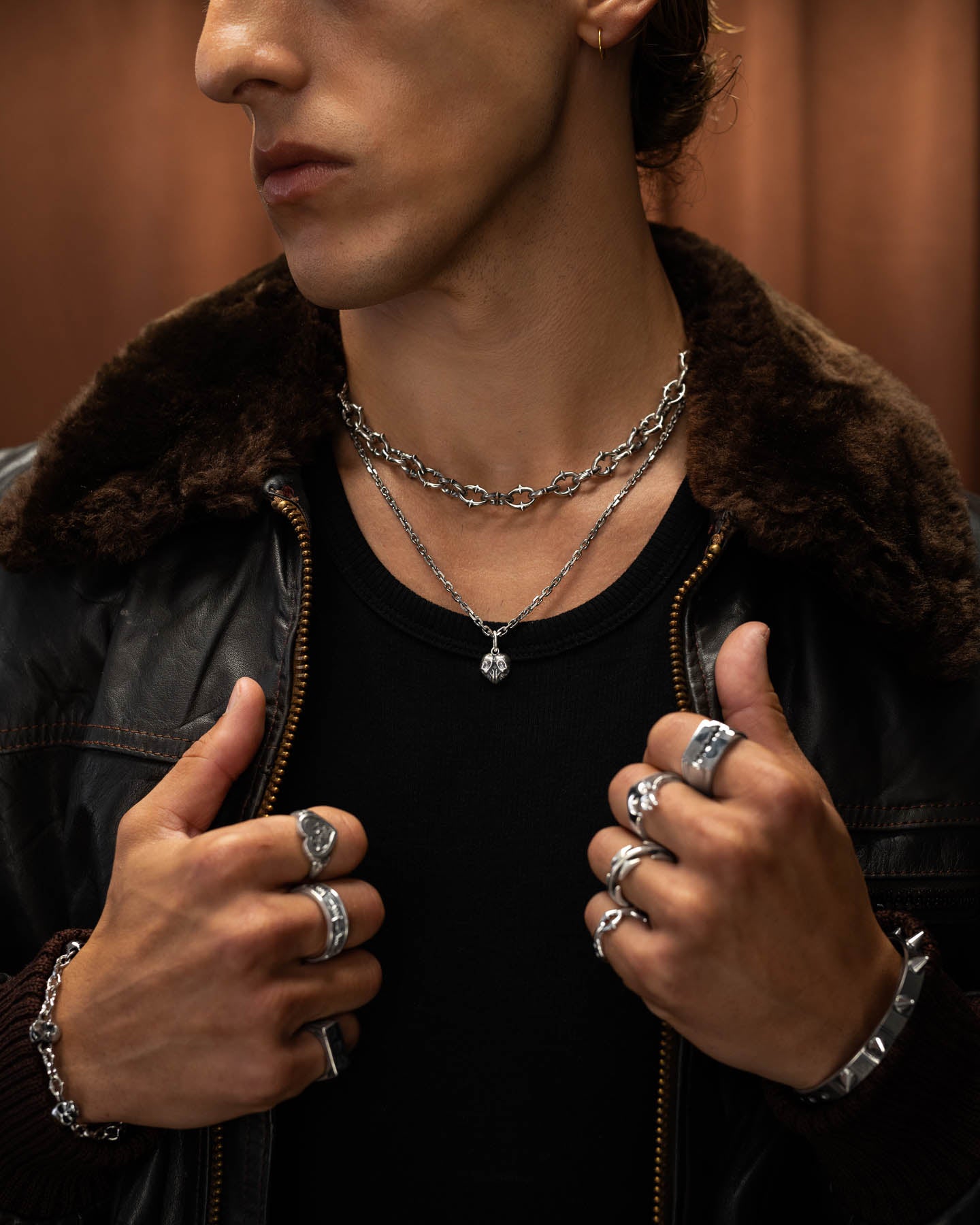A person in a dark shirt and brown shearling jacket, styled with rock jewelry—sterling silver rings, bracelets, and the 925 Sterling Silver Skull Heart Pendant—poses with hands near their chest against a softly blurred background.