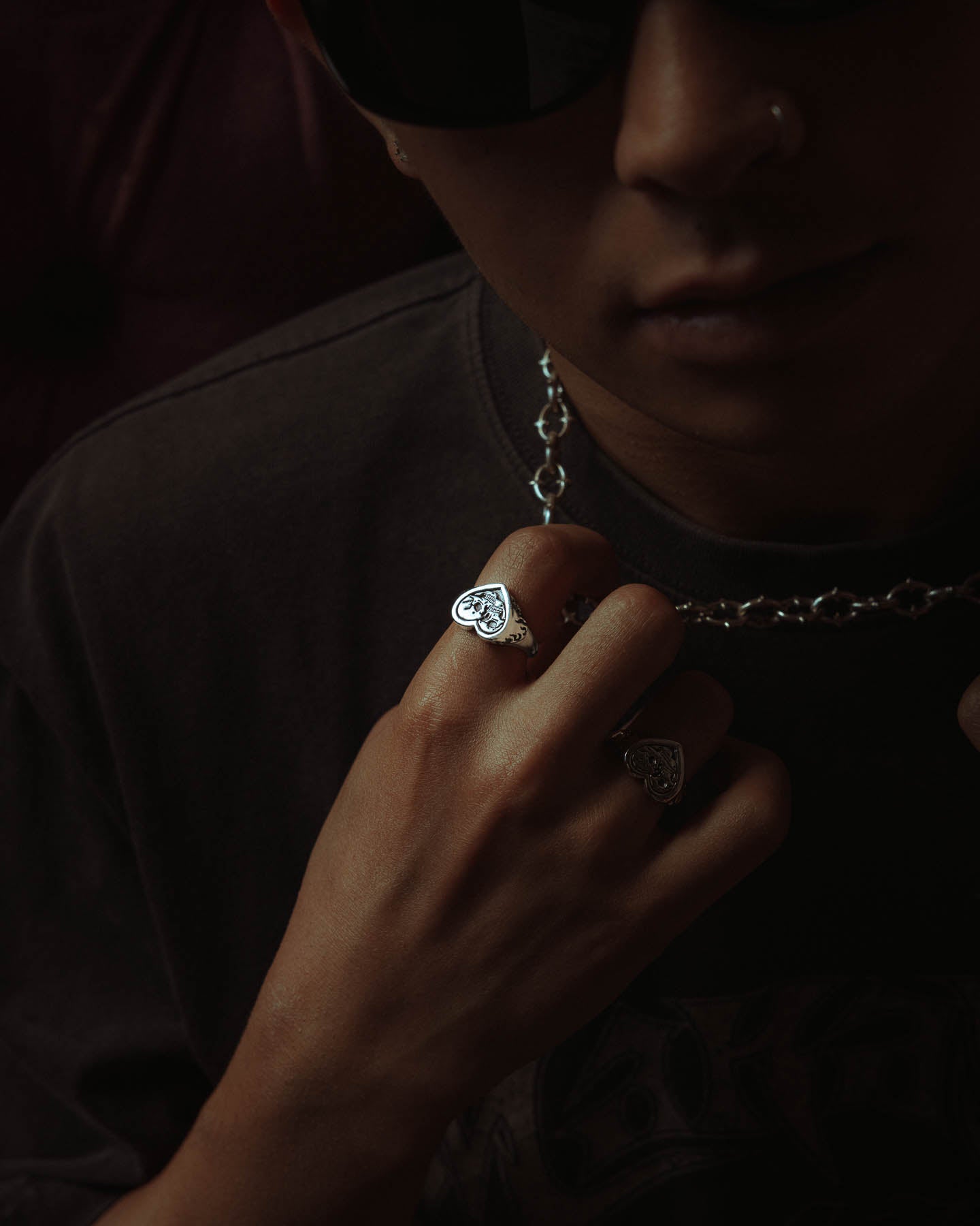 A person in dark sunglasses and a nose ring holds a chain necklace. Their hand is adorned with two handcrafted silver rings, including the 925 Sterling Silver Skull Heart Ring, while their face remains mostly in shadow.