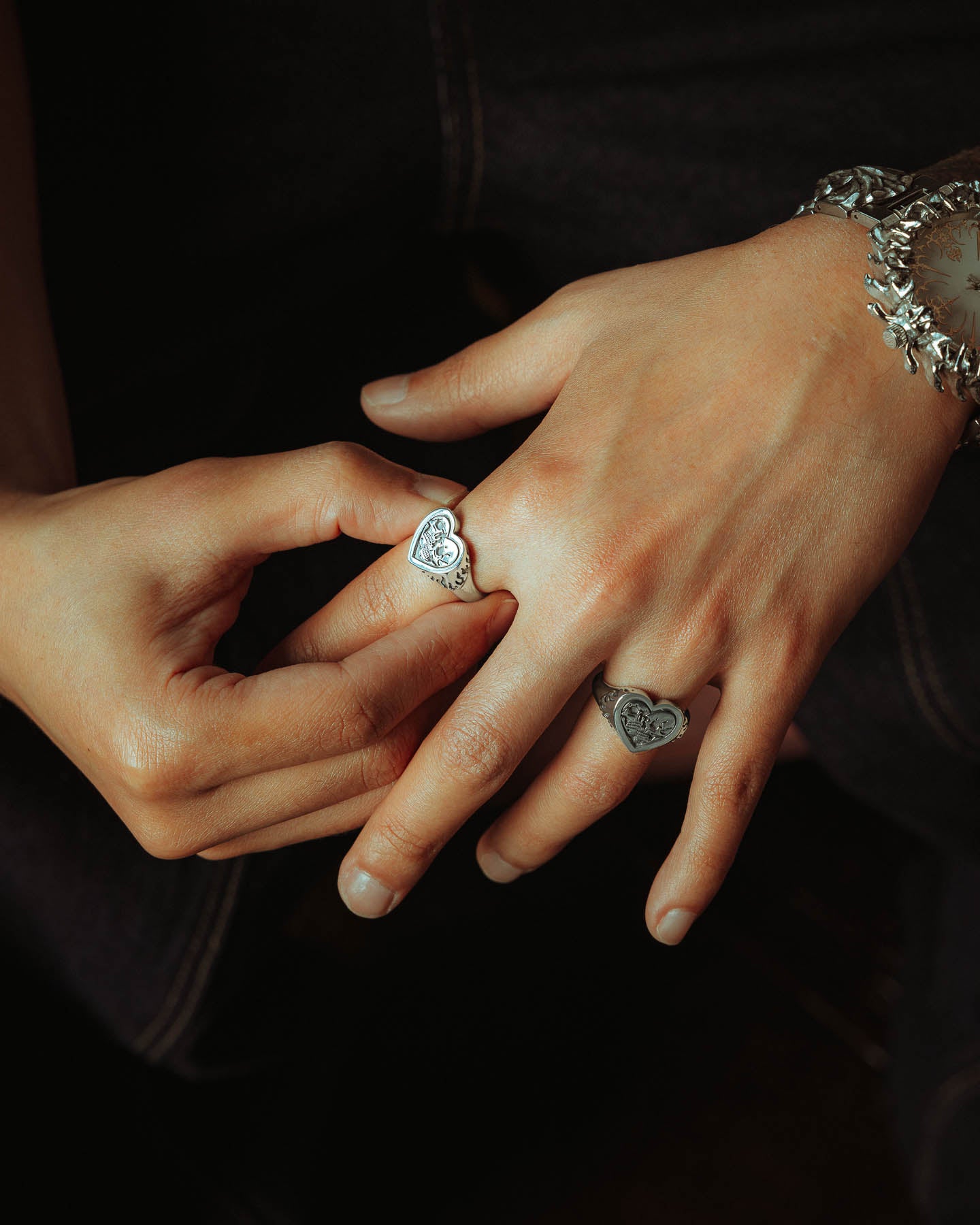 A close-up of two hands: one wears the 925 Sterling Silver Skull Heart Ring, while the other gently touches it, also adorned with a floral-patterned handcrafted silver bracelet and a watch against a dark background.