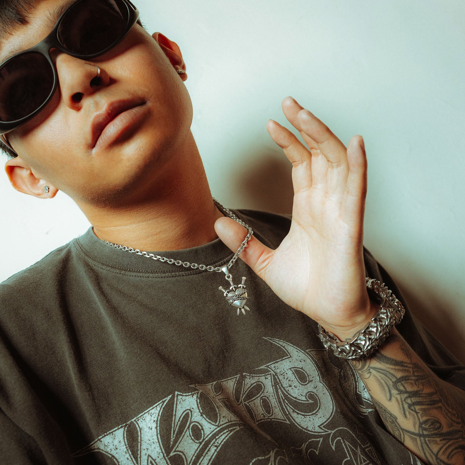 Silver heart pendant with 3 swords going through it and wrapped in chain on male model holding the chain up with glasses