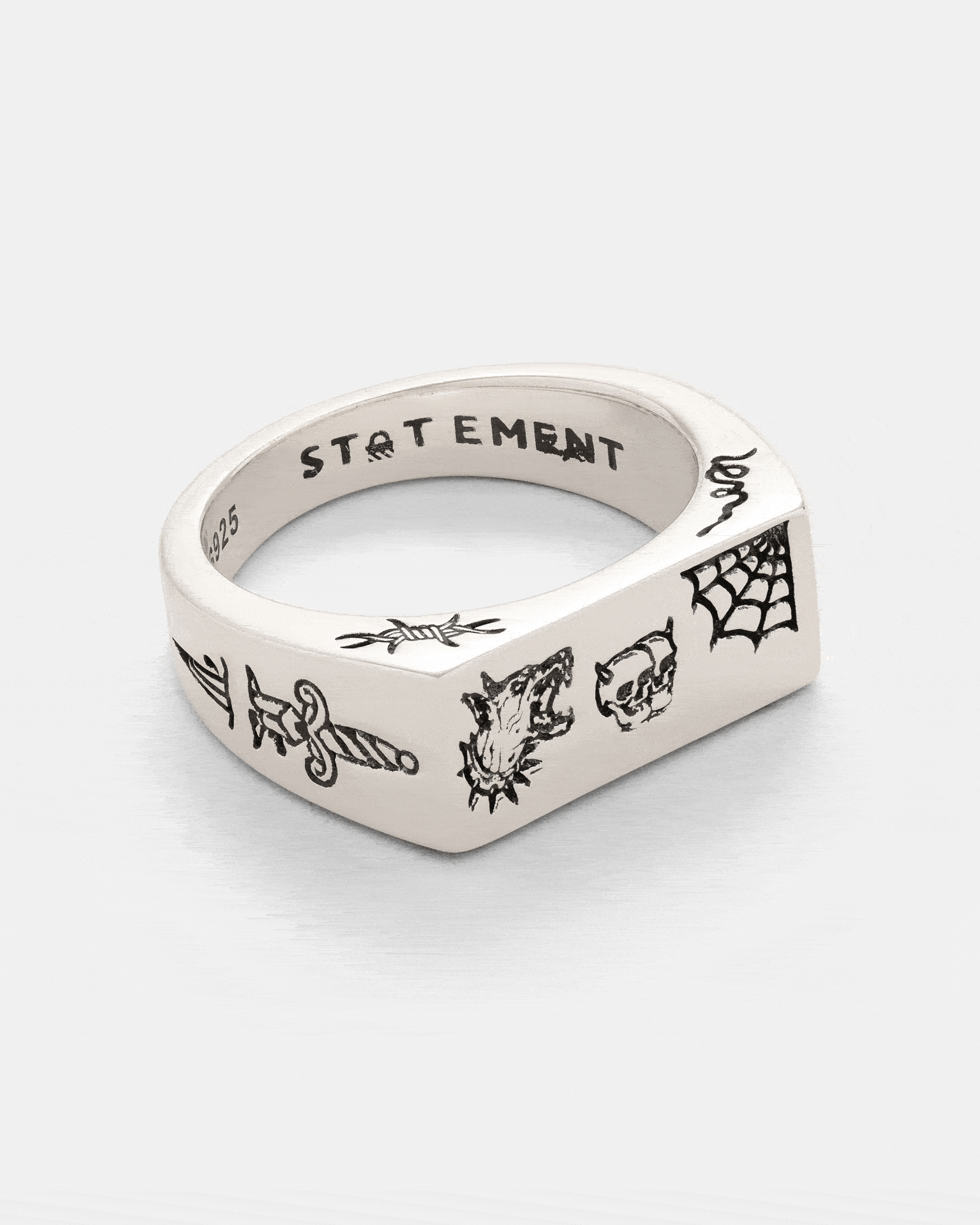 The 925 Sterling Silver Tattoo Signet Ring by Statement Collective showcases engraved black designs featuring a sword, wolf head, skull, and spider web. This bold ring displays a distinctive patchwork of motifs reminiscent of tattoo art. The inner band is stamped with “STATEMENT” and “925.”.