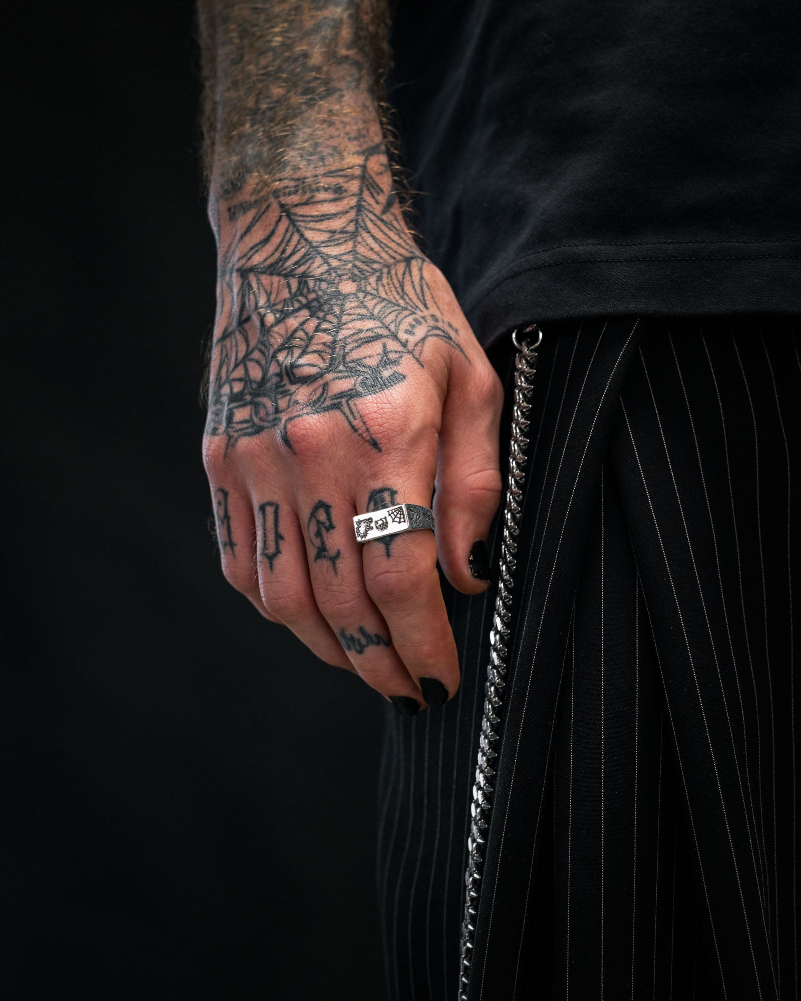 A tattooed hand with black nails wears the Statement Collective 925 Sterling Silver Tattoo Signet Ring, shown beside pinstriped pants with a chain. The tattoos feature a spider web and letters. The background is dark.