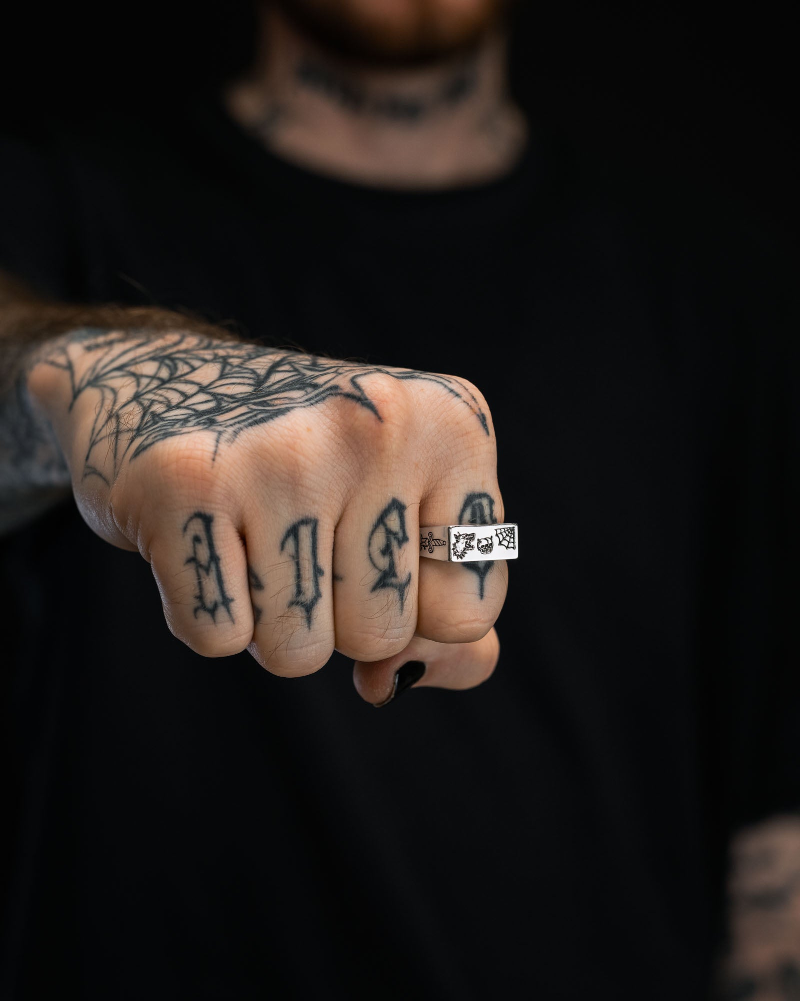 A tattooed fist extends toward the camera, showcasing the Statement Collective 925 Sterling Silver Tattoo Signet Ring, featuring engraved symbols. Gothic-style finger tattoos and a spiderweb design accent the look.