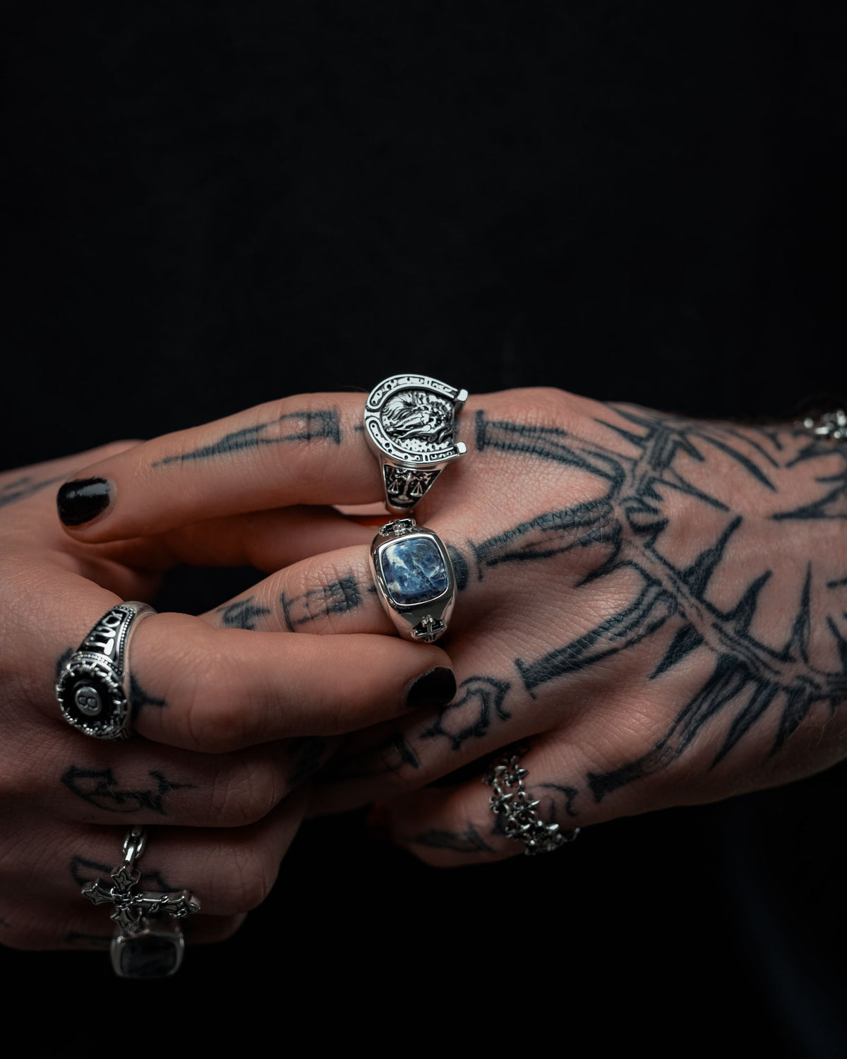 Tattooed hands with black nail polish showcase several silver rings by Statement Collective, including the 925 Sterling Silver Sodalite Stone Ring, a bold horse-inspired design, and an eye-catching razor wire ring. The dark background emphasizes the sterling silver jewelry and tattoo-themed details.
