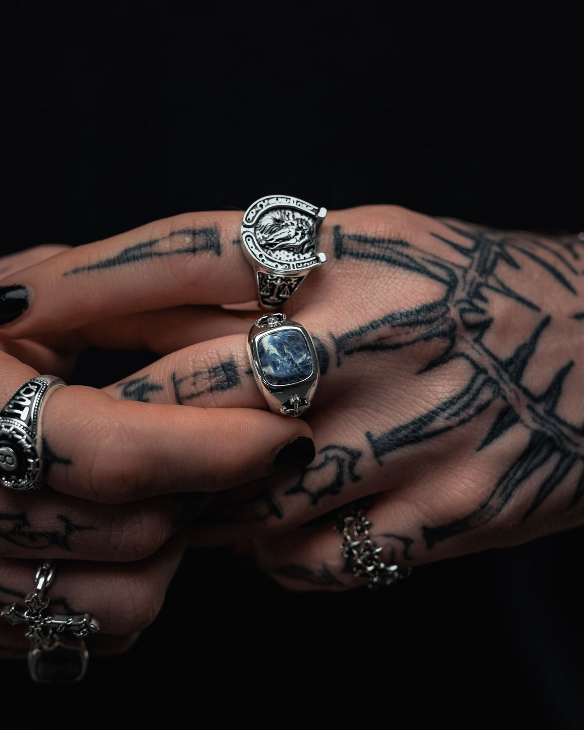 A close-up of tattooed fingers adorned with multiple pieces of sterling silver jewelry from Statement Collective, including the 925 Sterling Silver Sodalite Stone Ring featuring a striking blue sodalite stone, a bold razor wire ring, and another piece shaped like a horseshoe. The dark background accentuates the tattoo-inspired designs and intricate details of both the jewelry and tattoos.