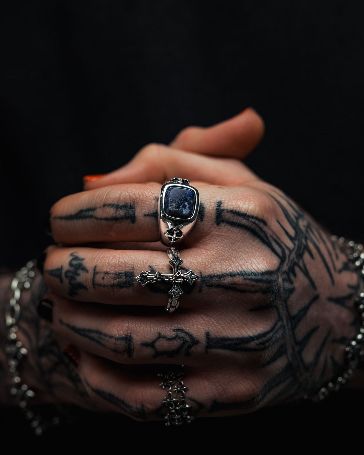 Tattooed hands clasped together, adorned with sterling silver jewelry from Statement Collective, including the 925 Sterling Silver Sodalite Stone Ring with a bold blue gemstone and a cross ring. A silver chain bracelet rests on the wrist—perfect for those drawn to tattoo-inspired accessories.