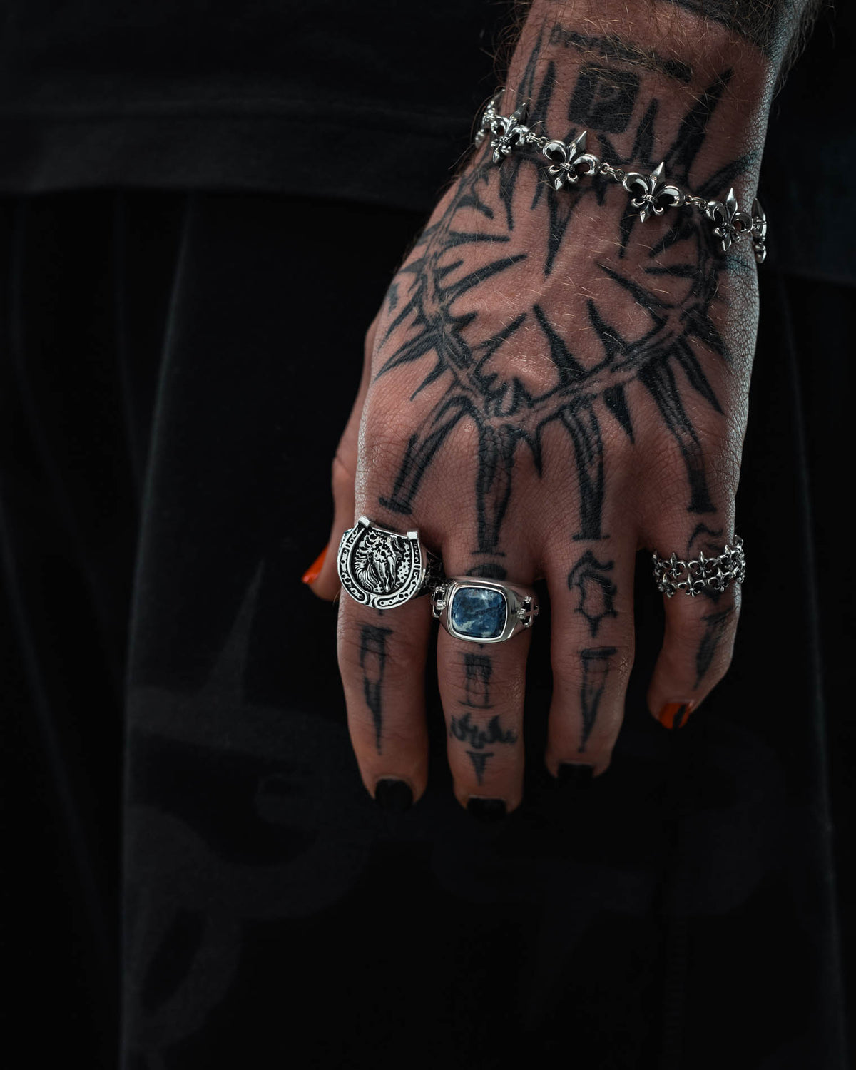 A tattooed hand with black-painted nails, adorned with macabre jewellery including Statement Collective’s 925 Sterling Silver Bronco Ring, gracefully wears a silver bracelet against dark clothing.