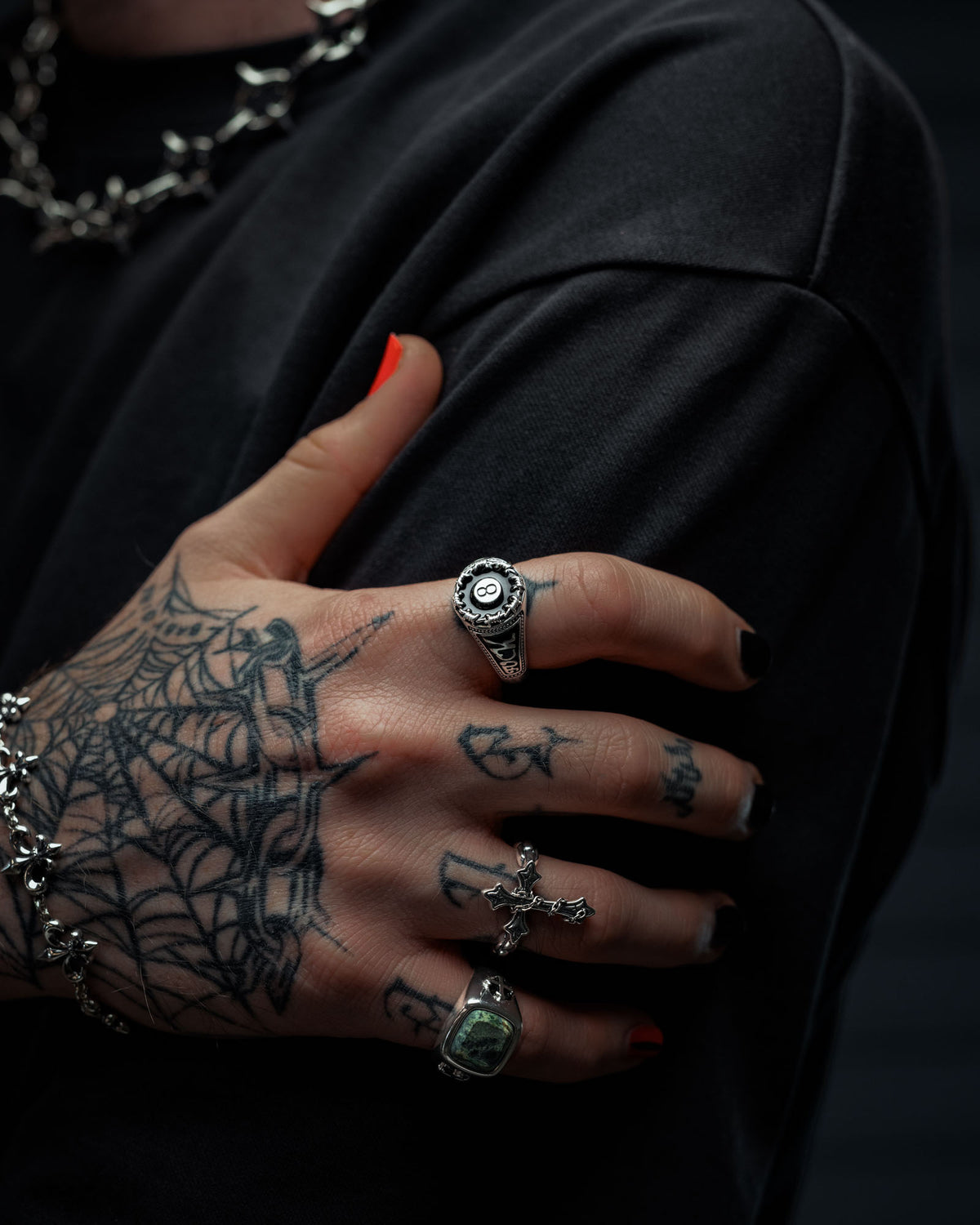 A close-up of a tattooed hand with black nail polish, adorned with multiple rings—including the eye-catching 925 Sterling Silver Lucky Shot Ring by Statement Collective—and a chain bracelet. The spiderweb tattoo on the hand rests against a black shirt, showcasing the bold style of tattoo-inspired jewelry.