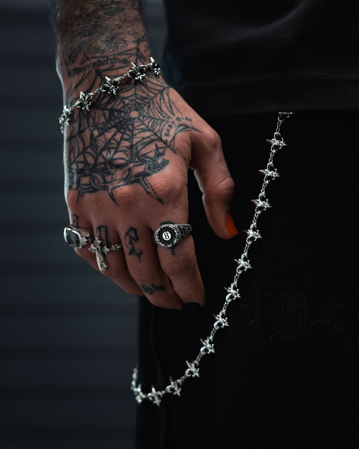 A tattooed hand adorned with multiple rings—including the eye-catching Statement Collective 925 Sterling Silver Lucky Shot Ring and a razor wire ring—as well as a bracelet, is displayed against a dark background. A chain with cross-shaped charms drapes from the wrist, perfectly capturing the bold essence of tattoo-inspired jewelry.