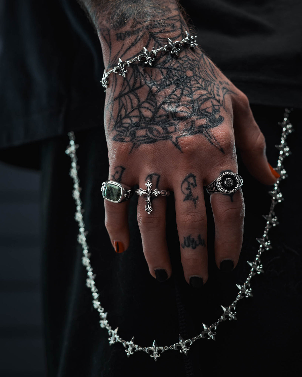 A tattooed hand with black nail polish is adorned with silver rings, including the Statement Collective 925 Sterling Silver “The Ruins” Chained Cross Ring, and holds a decorative chain. The hand also features a spider web tattoo and wears a bracelet with small cross charms.