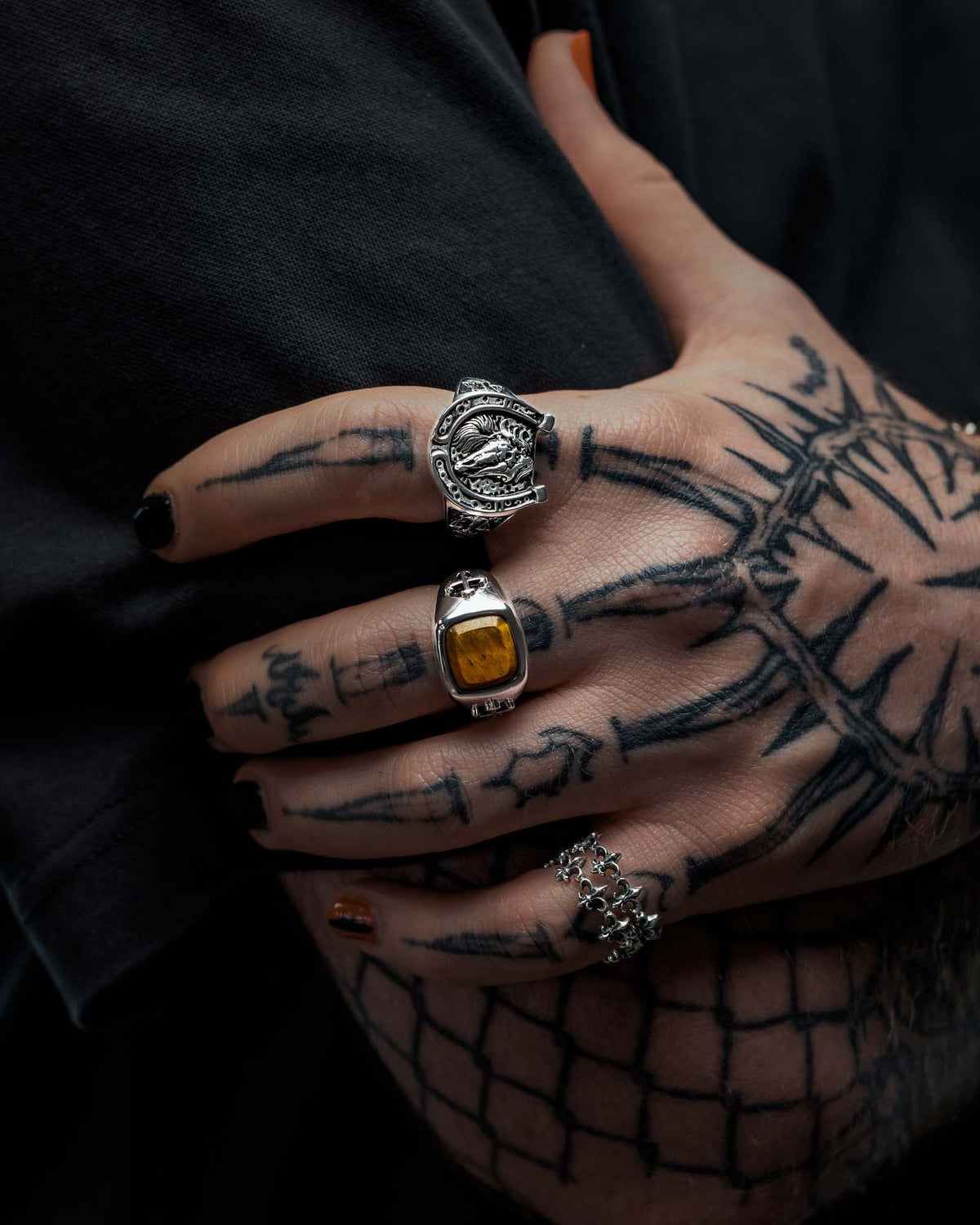 A tattooed hand with black nail polish showcases three silver rings, including the eye-catching 925 Sterling Silver Tigers Eye Stone Ring from Statement Collective. The geometric tattoos on the fingers and back of the hand enhance the bold, tattoo-inspired aesthetic of this jewelry look, all styled over a dark shirt.