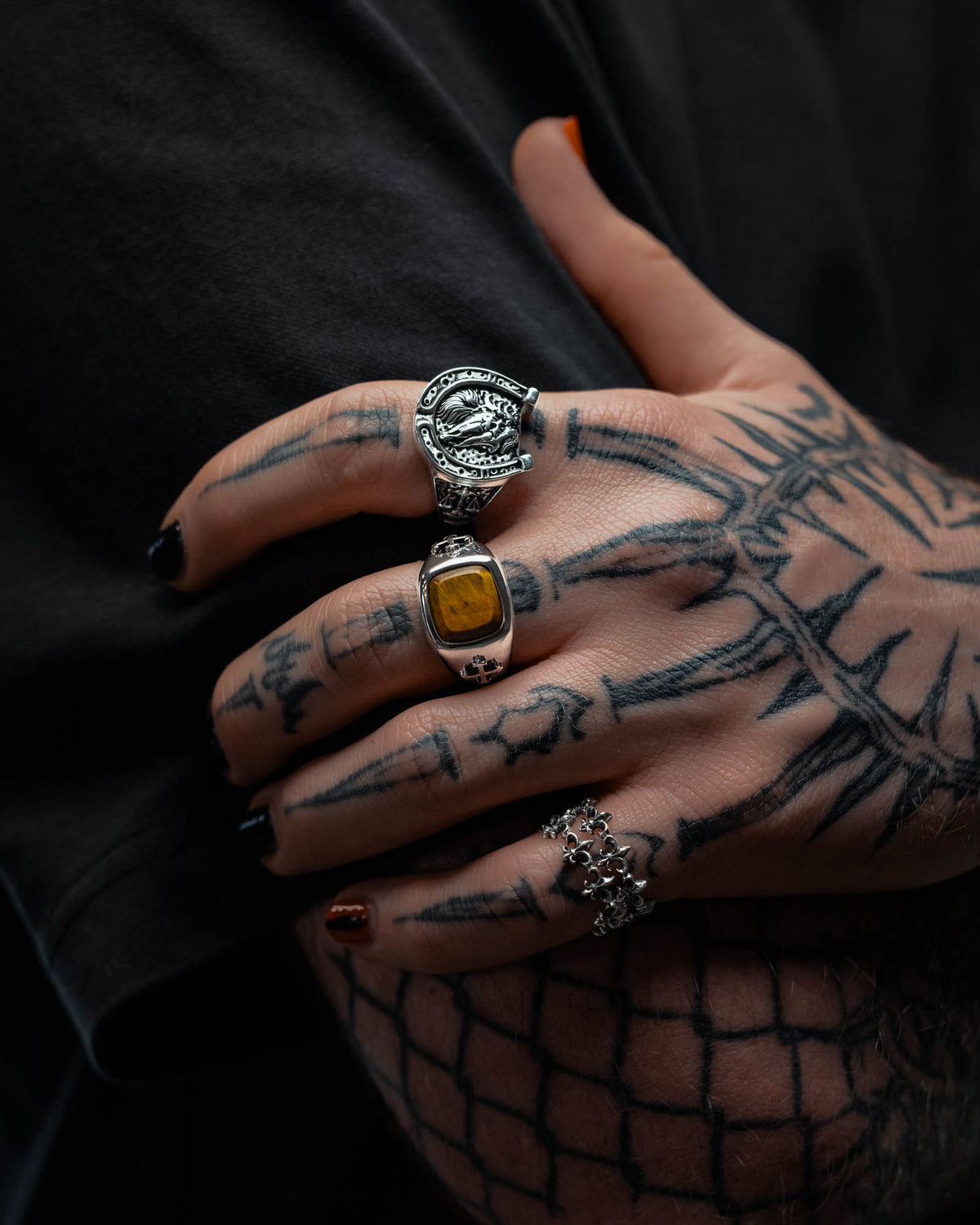 A tattooed hand with black-painted nails showcases three unique rings: the 925 Sterling Silver Tigers Eye Stone Ring by Statement Collective, a large sterling silver ring with an emblem, and a silver ring adorned with small skulls. Drawing inspiration from tattoo-inspired jewelry, the look is finished off with the individual’s dark shirt.