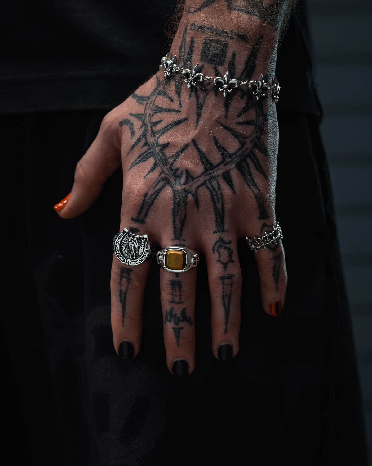 A tattooed hand with black nail polish and silver rings, including the eye-catching 925 Sterling Silver Tigers Eye Stone Ring by Statement Collective. The hand also flaunts a razor wire ring inspired by tattoo art, a sleek silver bracelet, and one fingernail painted orange for an edgy pop of color.