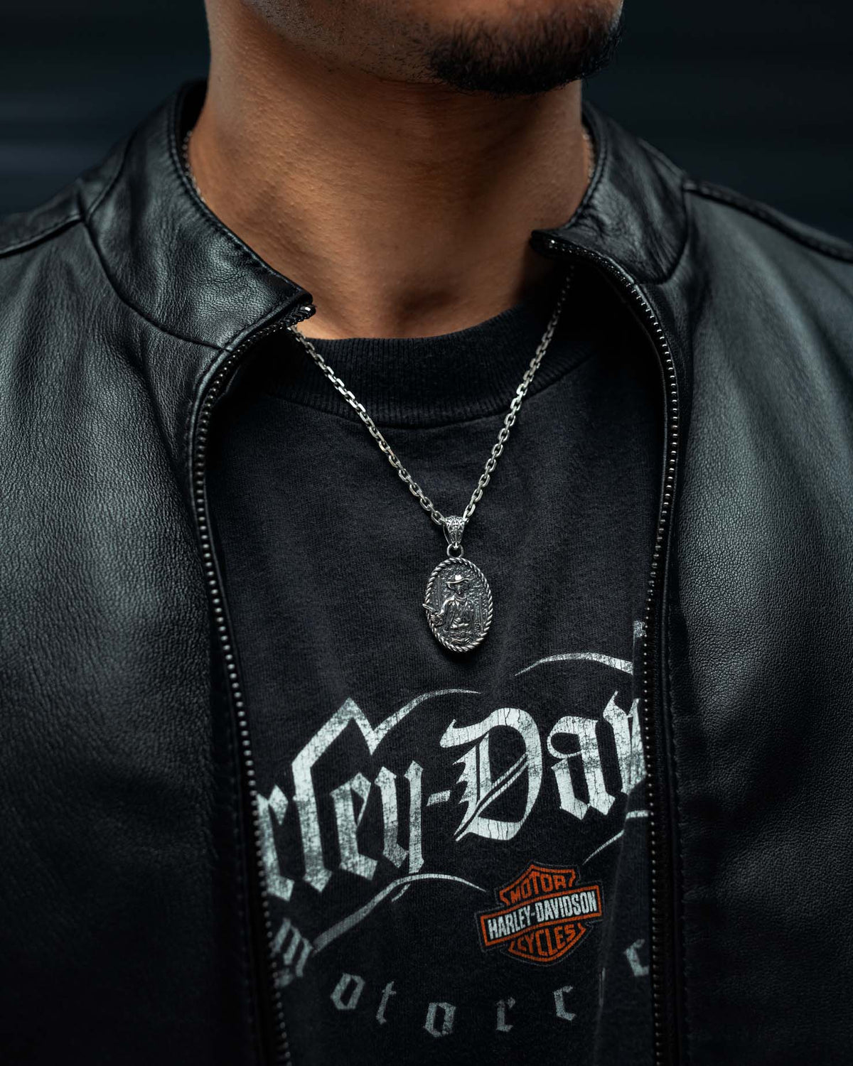 A person wearing a black leather jacket, a Harley-Davidson t-shirt, and the Statement Collective 925 Sterling Silver “Big Iron” Pendant necklace. Only the lower face, neck, and upper chest are visible.