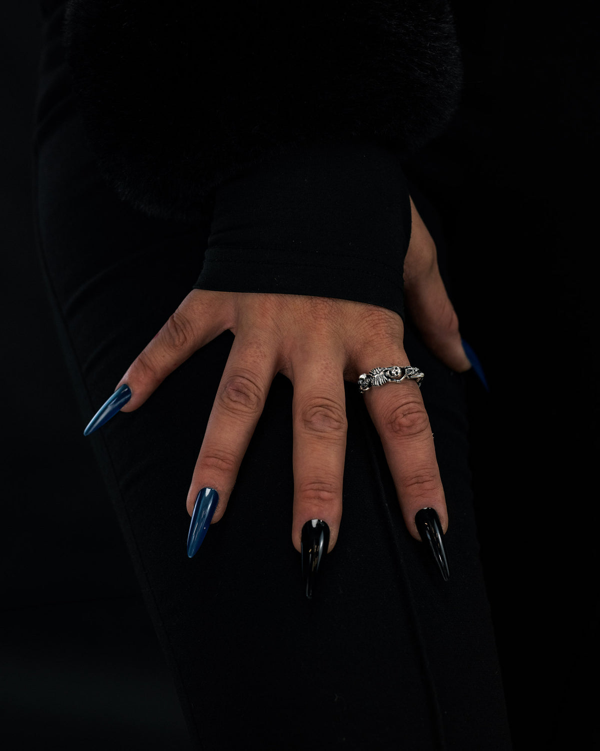 A hand with long, pointed black and blue nails rests on a thigh. The person wears the Statement Collective 925 Sterling Silver Breaking Wheel Ring with its decorative design and is dressed in black clothing featuring a plush, textured sleeve.